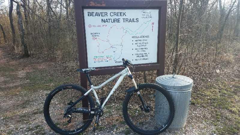 A mountain bike leaning against a sign that reads "Beaver Creek Nature Trails," which includes a map of the trail and a list of regulations for the area. A trash can is visible next to the sign, and the surrounding area features gravel paths and trees. The scene suggests a recreational outdoor setting for biking and nature exploration. Orange Loop mountain bike trail.