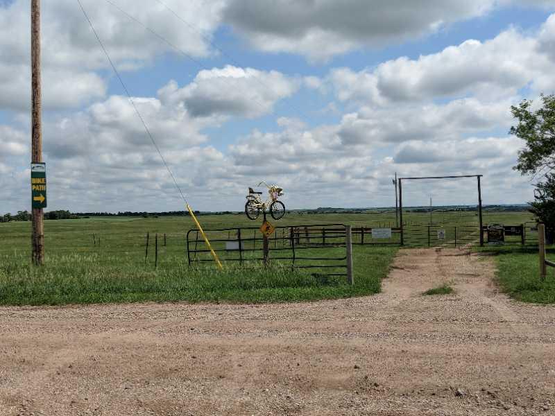 A gravel road leads to an open gate, bordered by green grass and fields under a partly cloudy sky. A signpost indicates a bike path with an arrow to the left, and a yellow bicycle hangs on the gate, decorated with flowers. Black Butte Adventures mountain bike trail.