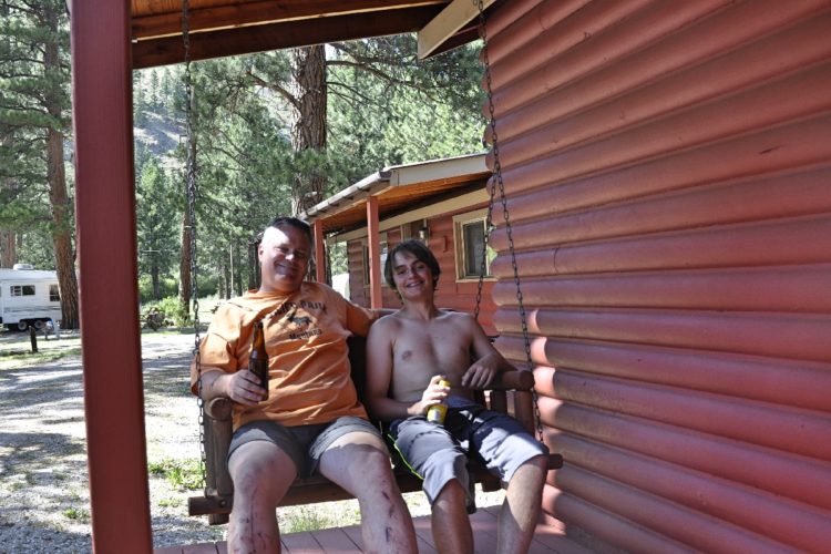 Two individuals sit together on a wooden swing outside a rustic cabin. One person is wearing an orange shirt and shorts, holding a drink, while the other is shirtless in shorts, also holding a beverage. They are smiling and enjoying the outdoors, with trees and a parked trailer visible in the background. The cabin has a log-style exterior painted in red.