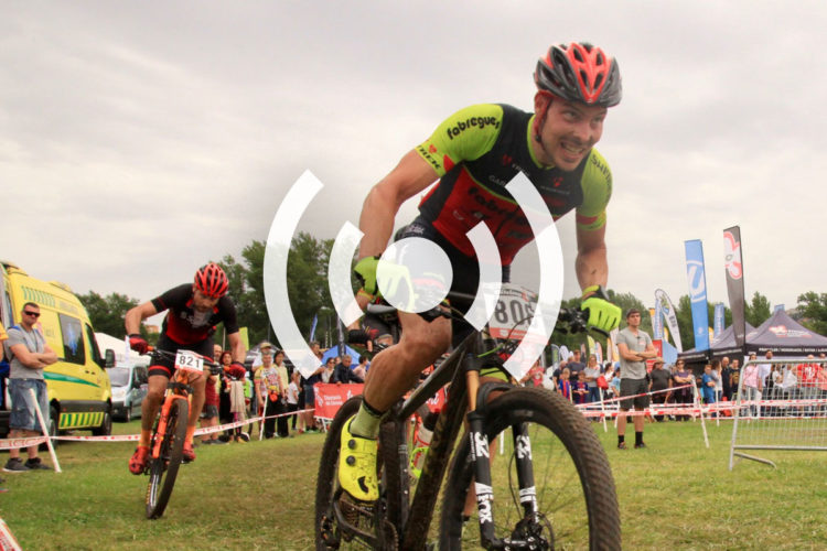 Two mountain bikers competing in a race, with one rider in the foreground smiling and focused as he pedals on a dirt path. The other rider is slightly behind him, leaning forward in an athletic position. A crowd watches in the background, with tents and banners visible, indicating a lively event atmosphere.