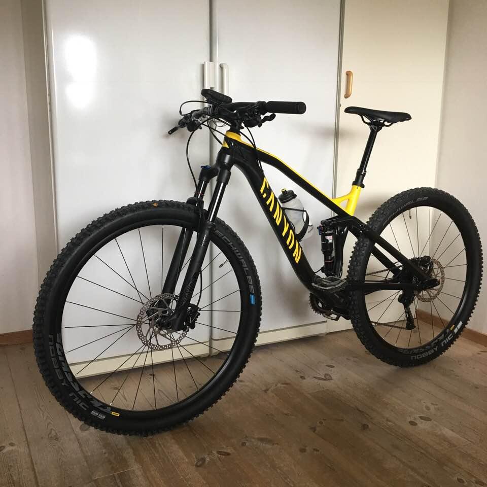 Canyon Neuron: A black and yellow mountain bike is leaning against a white cabinet in a room with wooden flooring. The bike features wide tires, a front suspension fork, and disc brakes. A water bottle is mounted in the frame. The background includes a plain wall and a closed closet.