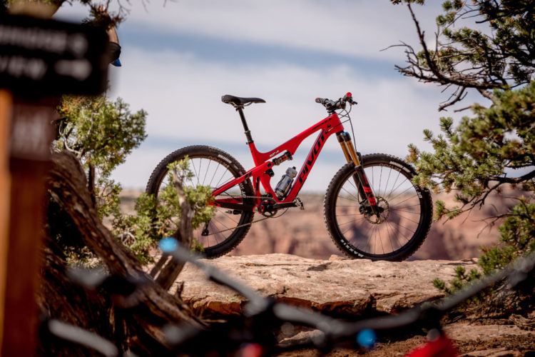 A vibrant red mountain bike is positioned on a rocky outcrop, surrounded by greenery and natural scenery. In the background, a scenic landscape extends into the distance under a partly cloudy sky. A wooden trail sign is partially visible on the left.