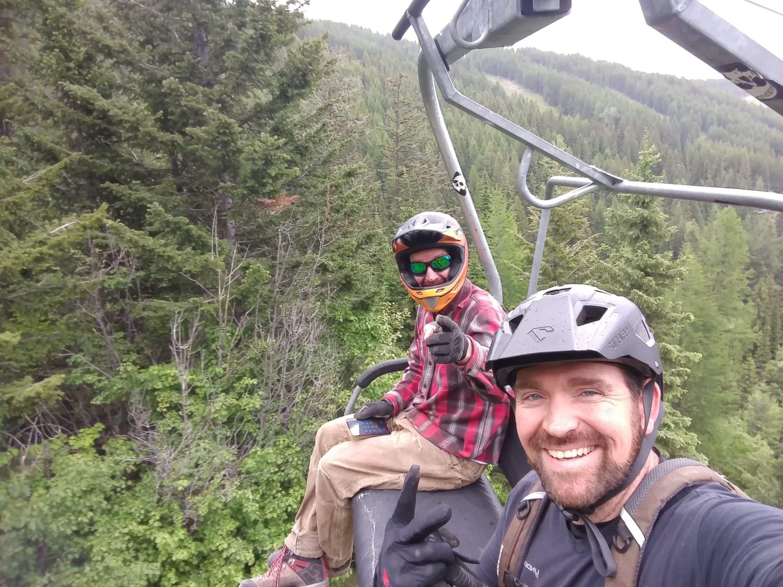 Two mountain bikers sitting on a ski lift, surrounded by lush green trees. One biker is smiling and giving a thumbs-up, while the other is pointing and wearing sunglasses and a helmet. The background features a mountainous landscape with varied foliage. Whitefish Mountain Resort mountain bike trail.