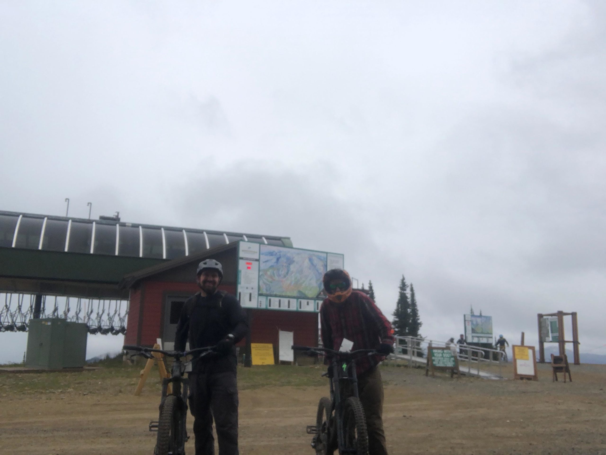 Two mountain bikers wearing helmets and riding gear stand next to their bicycles at a mountain resort. In the background, there is a ski lift station and a map of the trails. The sky is overcast, indicating a cloudy day. Whitefish Mountain Resort mountain bike trail.