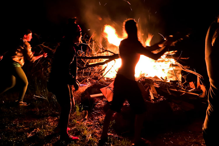A group of people gathered around a large bonfire at night, with flames and sparks illuminating the scene. Some individuals are actively adding branches to the fire, while others are seen in motion, creating a lively atmosphere. The dark surroundings contrast with the bright firelight, enhancing the warmth and energy of the gathering.
