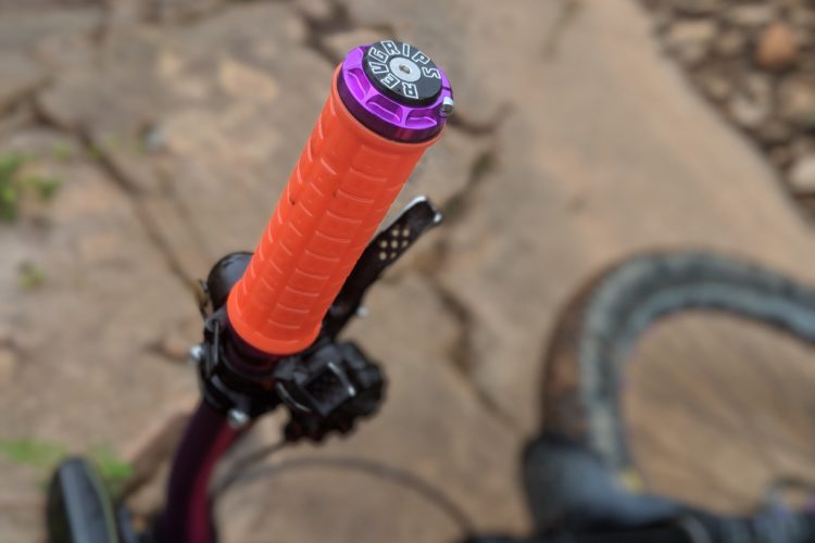 Close-up of a colorful bicycle handlebar grip, featuring an orange grip with textured ridges and a purple top. The background shows a rocky surface, suggesting an outdoor or trail setting.