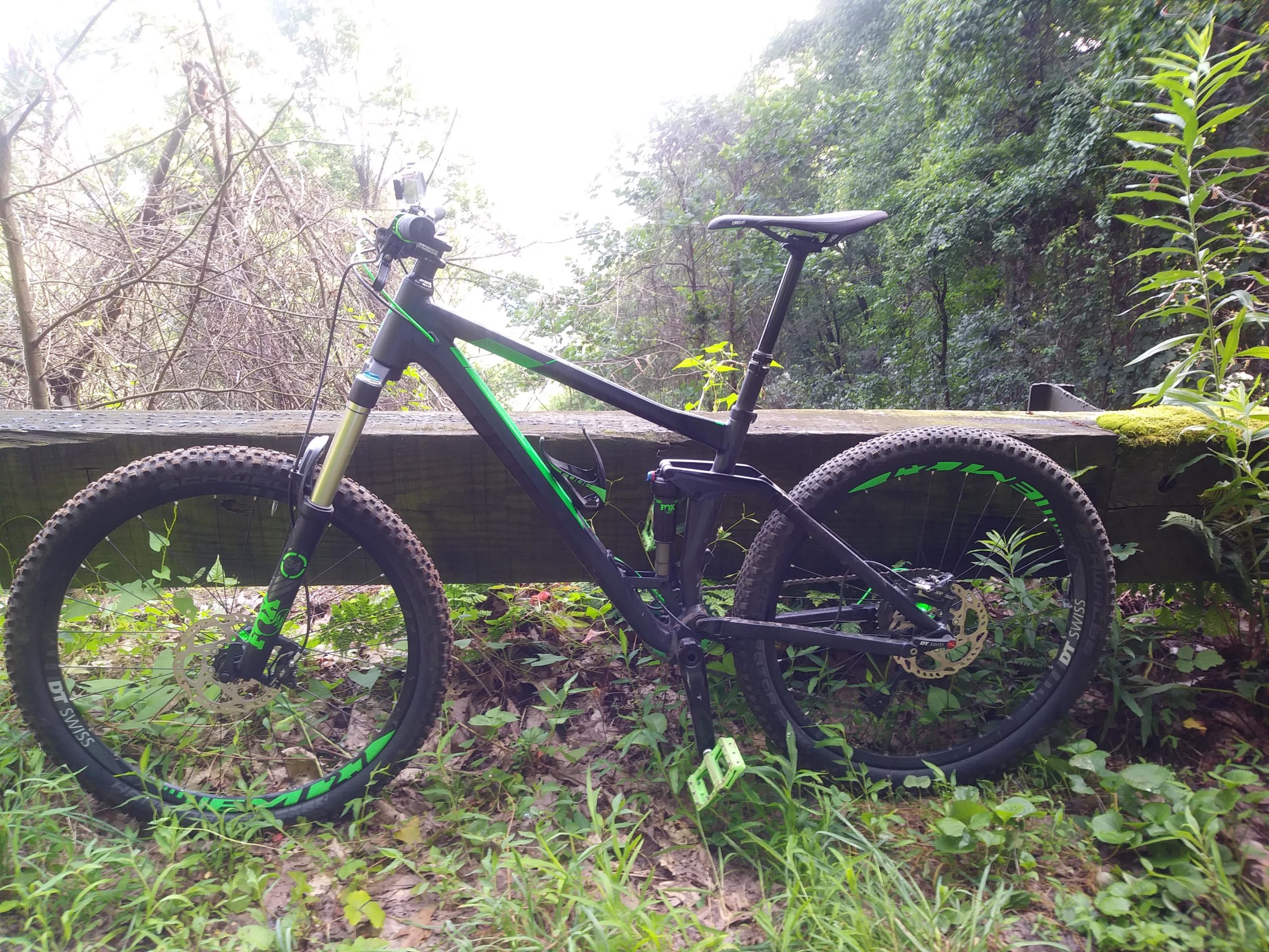 A black mountain bike with green accents is parked beside a wooden railing in a lush, wooded area. The bike features rugged tires and a suspension fork, with foliage and tall grass surrounding it. In the background, dense trees add to the natural scenery. Bent Creek mountain bike trail.