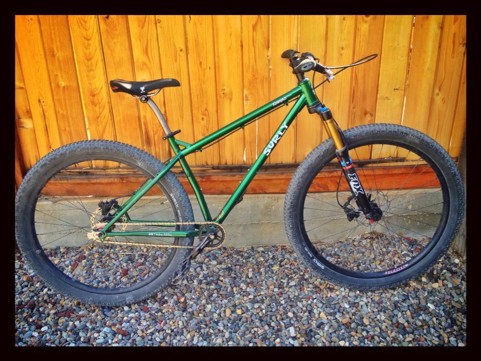 Surly Krampus: A green mountain bike with wide, knobby tires is leaning against a wooden fence. The bike features a sturdy frame, a stylish black seat, and a gold front fork, indicating it is designed for off-road use. The ground is covered with small pebbles, complementing the rustic background.