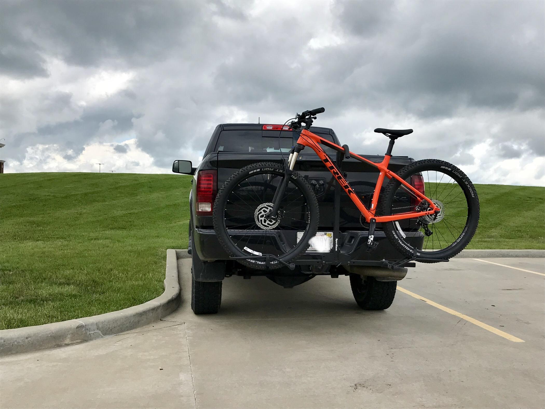 A black truck parked in a lot with an orange mountain bike mounted on a rear bike rack, set against a backdrop of cloudy skies and a grassy hill.