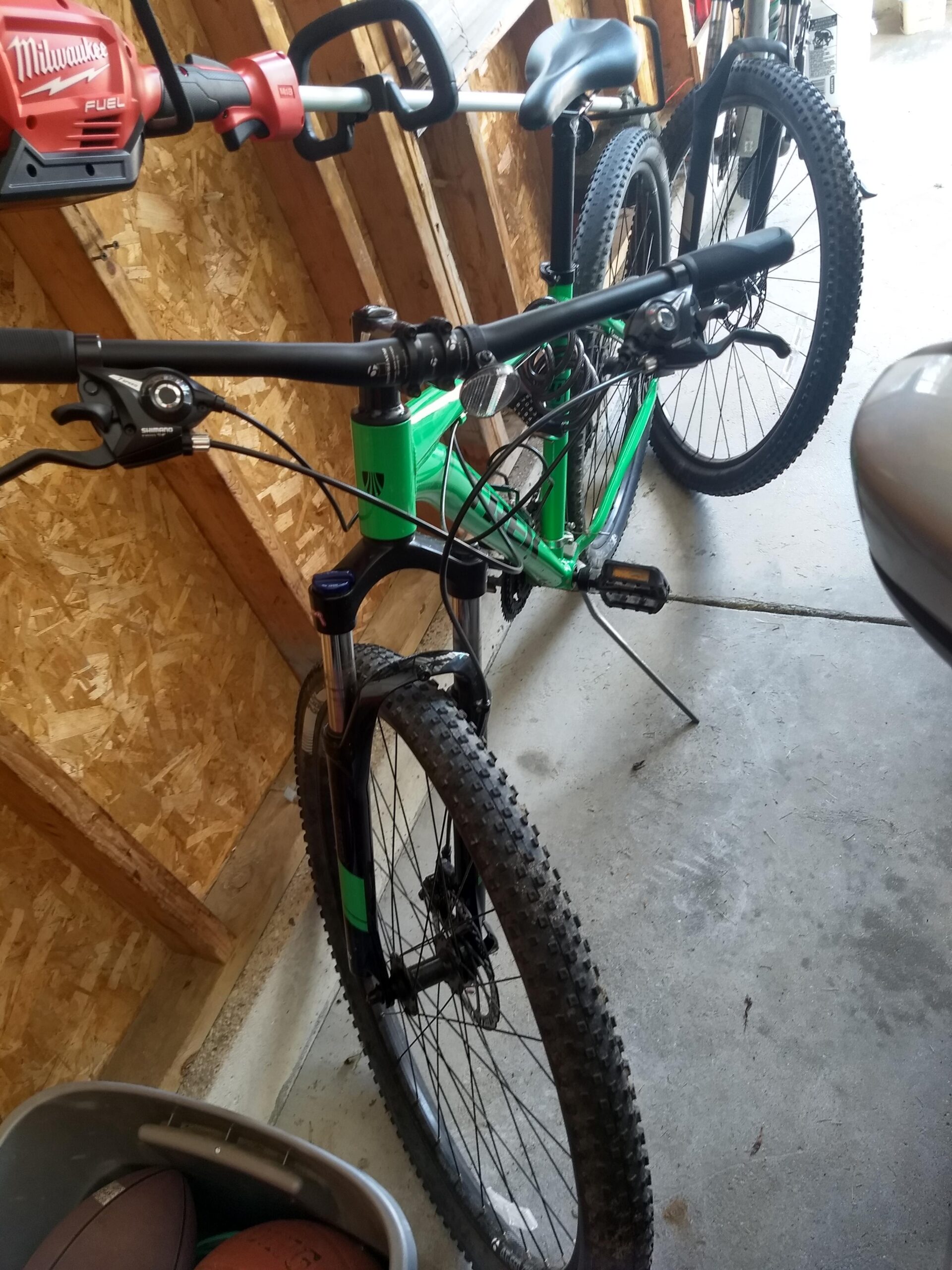 Trek Marlin 5 29er: A green mountain bike positioned in a garage, partially obscured by a lawn tool mounted above it. The bike has thick, knobby tires and a black fork. Nearby, a plastic bin contains sports balls. The garage features wooden walls and a concrete floor.