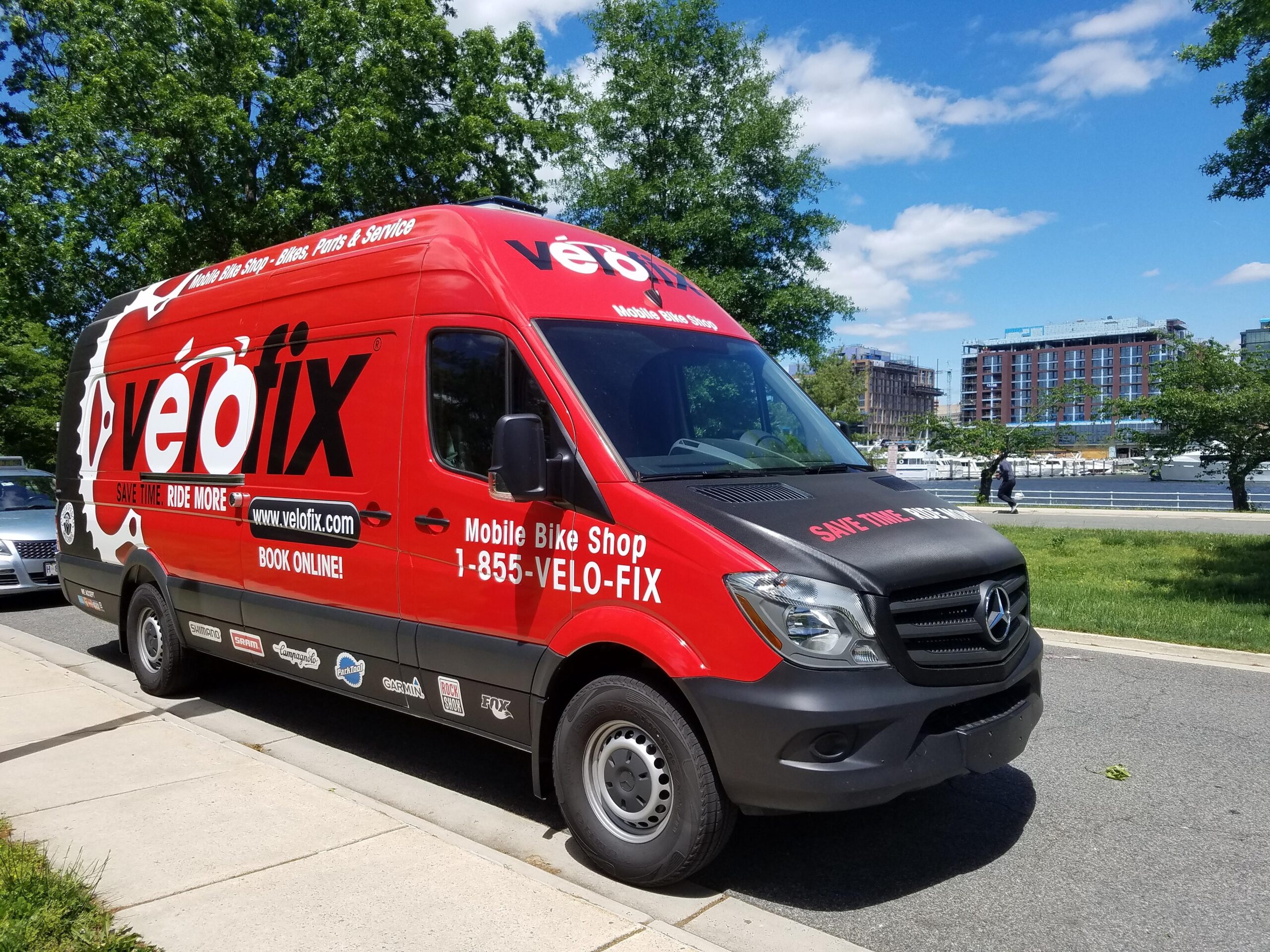 Red mobile bike shop van parked alongside a sidewalk, featuring the logo "velofix" prominently on the side. The vehicle also displays text promoting bike repair services, website, and contact number. In the background, there are trees, a body of water, and buildings under a clear blue sky.
