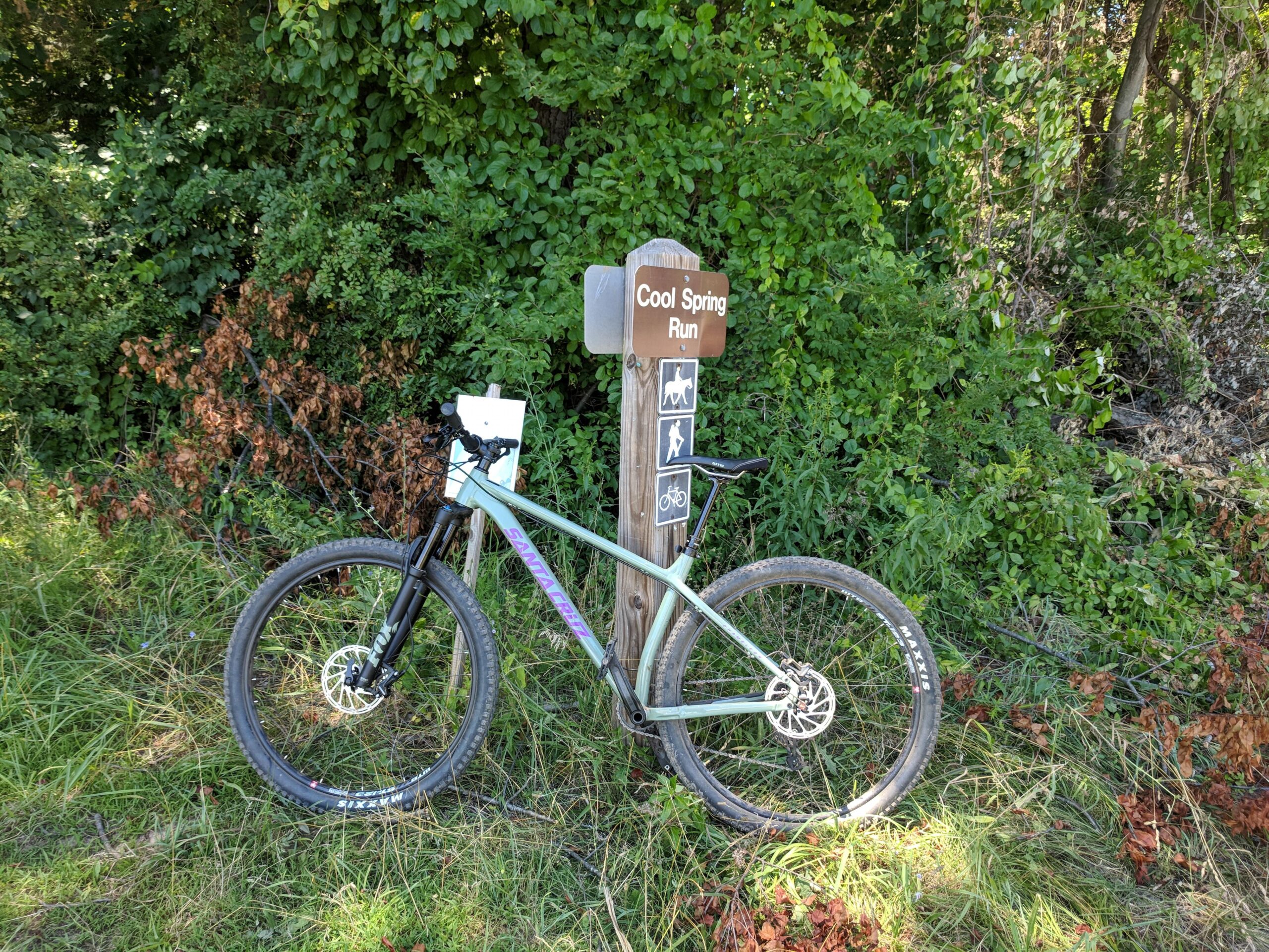 Santa Cruz Chameleon: A mountain bike parked next to a trail sign that reads "Cool Spring Run," surrounded by lush green foliage and grass. The sign includes icons for hiking, horseback riding, and biking.