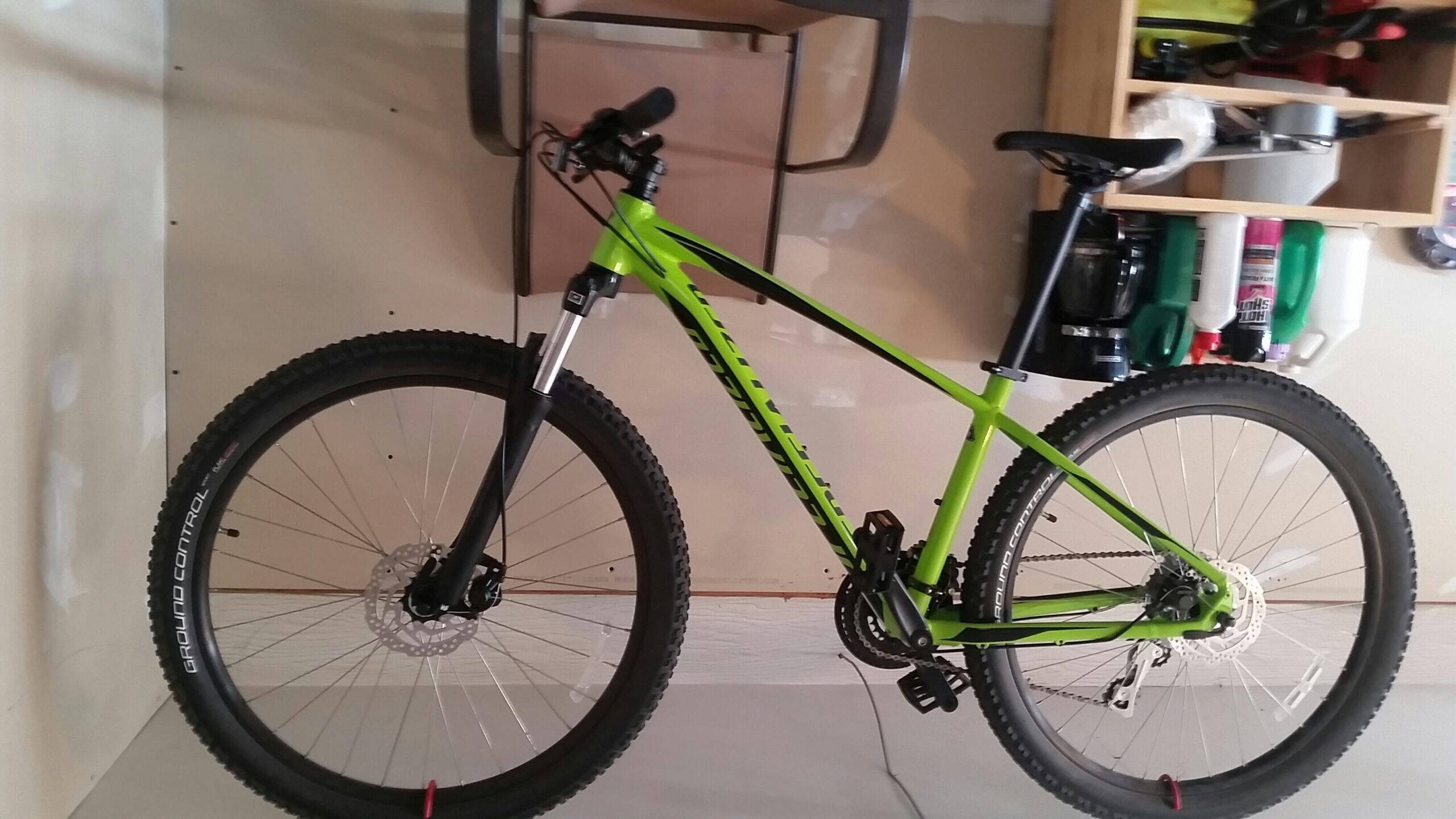 Specialized Pitch: A vibrant green mountain bike leaning against a light-colored wall in a garage, with a visible disc brake and knobby tires. The background features shelves with various tools and containers.