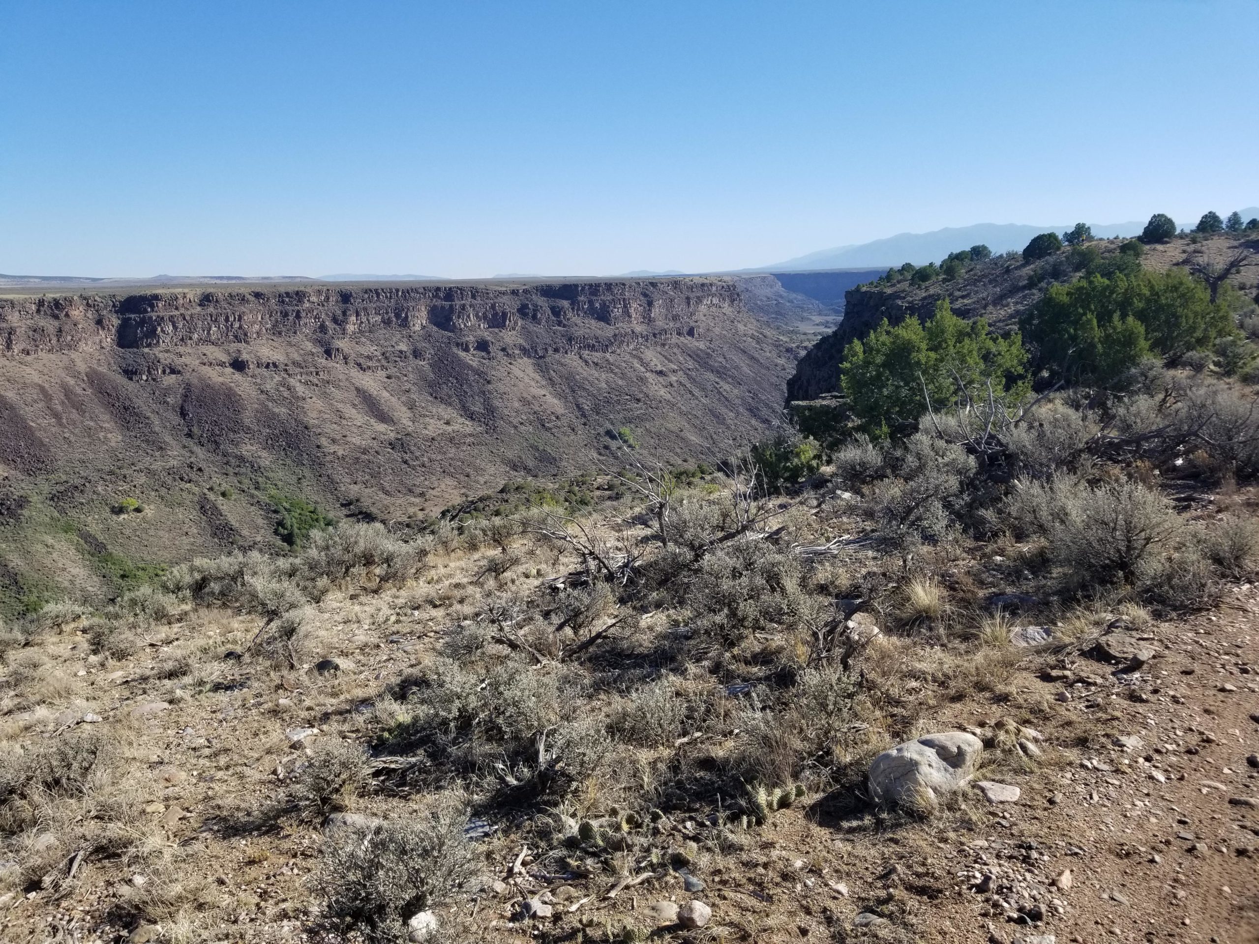 A panoramic view of a rugged canyon landscape, featuring steep cliffs and a vast, dry valley. The foreground includes rocky terrain and sparse shrubs, while the background showcases layered cliffs under a clear blue sky. Taos Valley Overlook mountain bike trail.