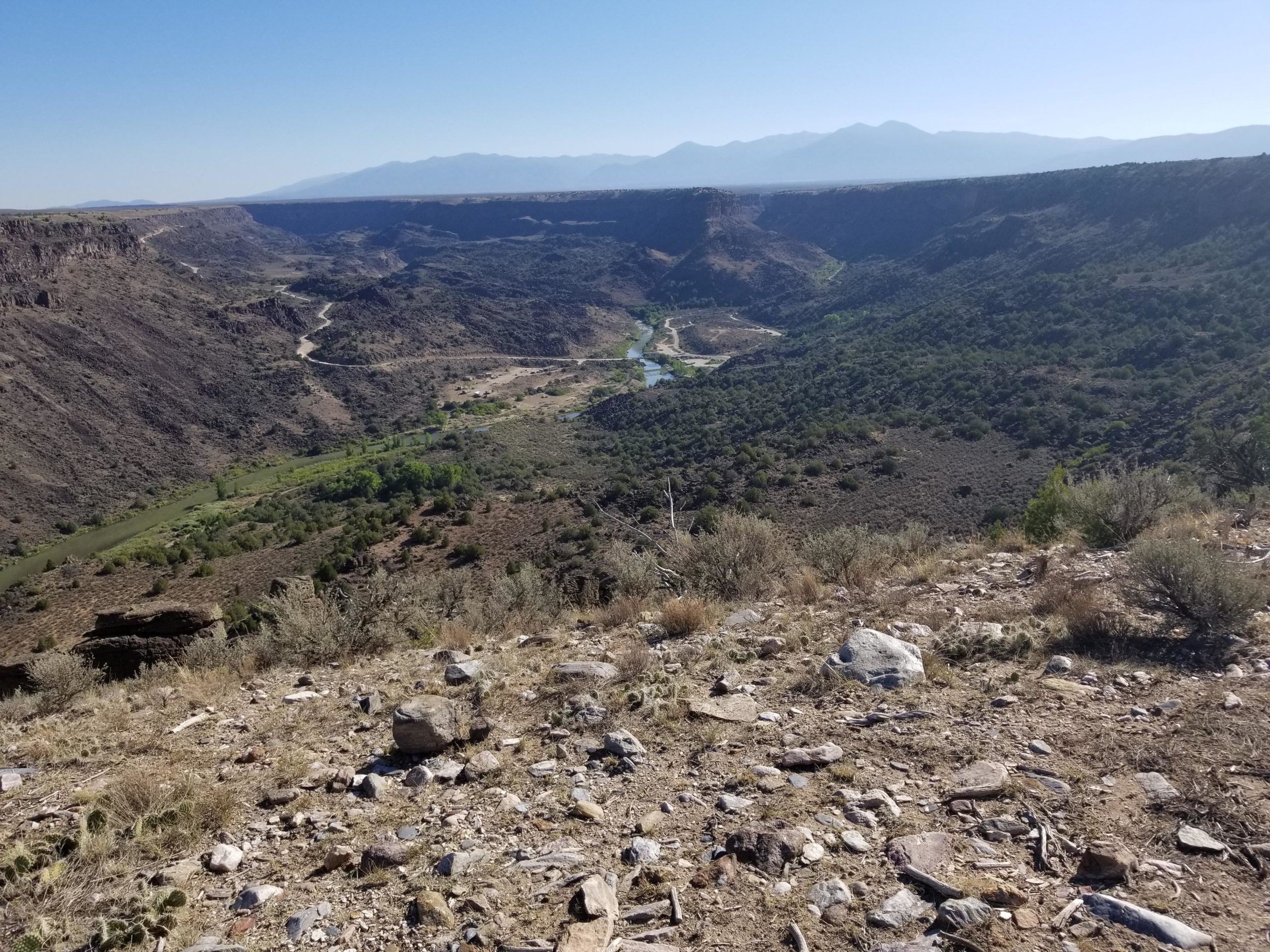 A panoramic view of a rugged canyon landscape, featuring steep cliffs and a winding river below. The foreground includes rocky terrain and sparse vegetation, while the background showcases distant mountains under a clear blue sky. Taos Valley Overlook mountain bike trail.