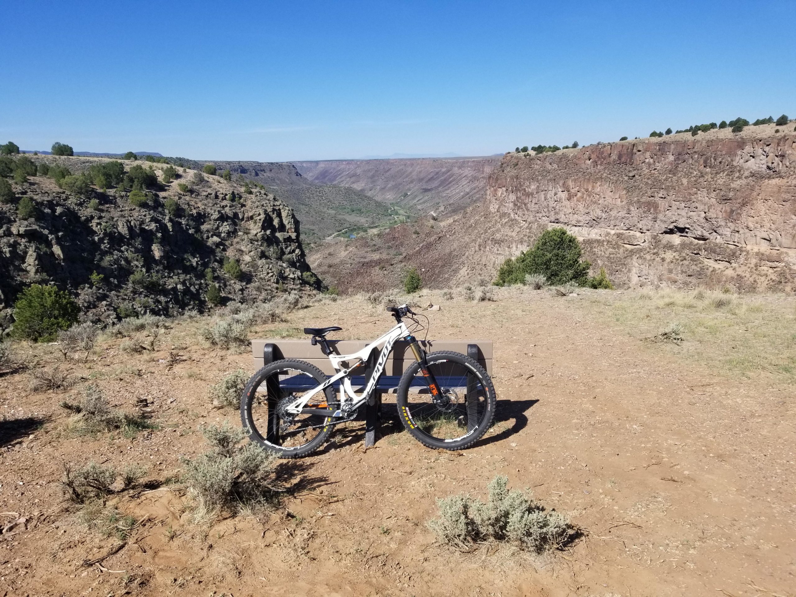 A mountain bike parked on a dirt trail overlooking a canyon landscape with greenery and rocky cliffs under a clear blue sky. Taos Valley Overlook mountain bike trail.