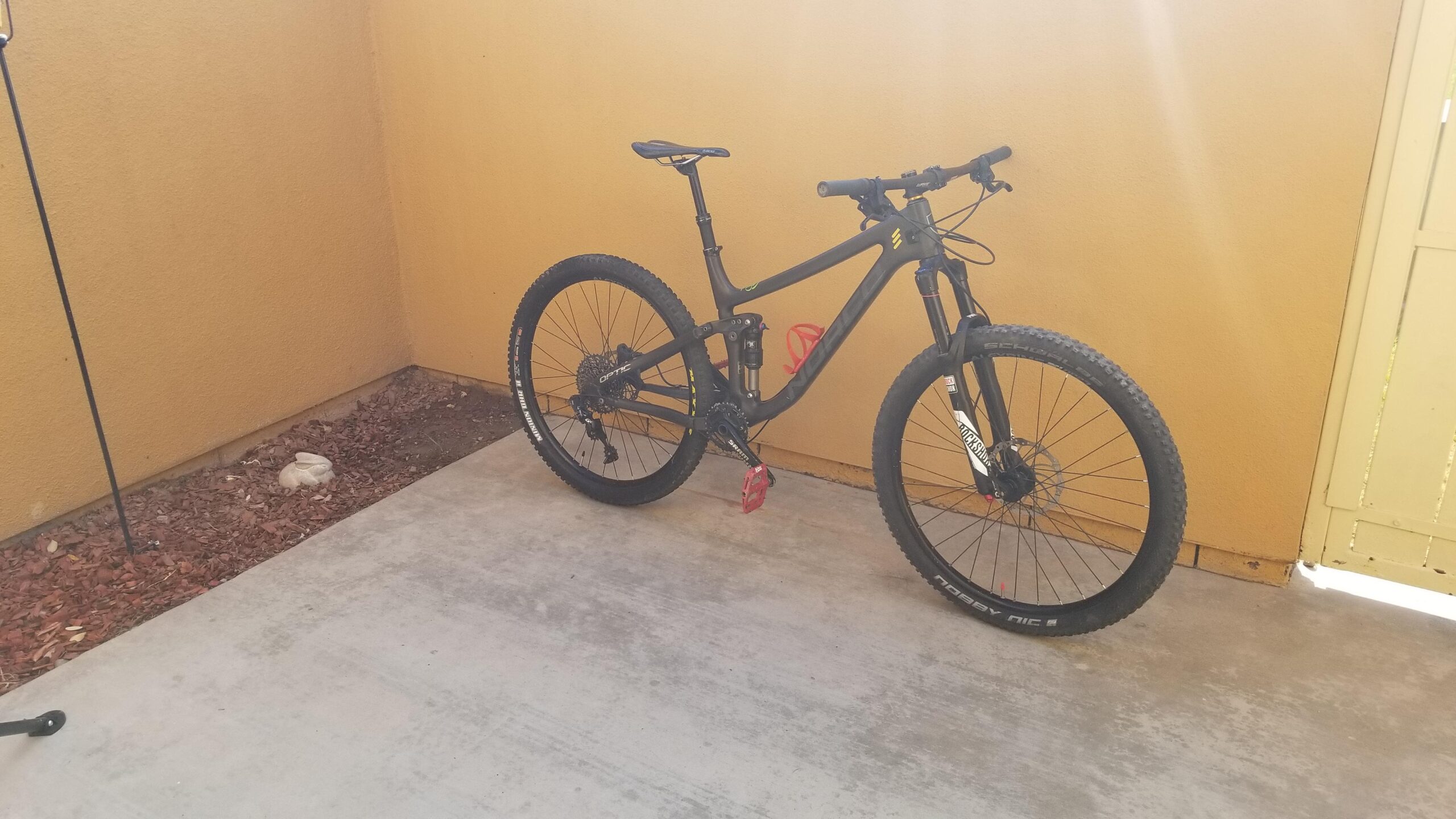 Norco Optic: A black mountain bike with colorful accents is parked on a concrete surface against a yellow wall. The bike features wide tires and a prominent front suspension, indicating it