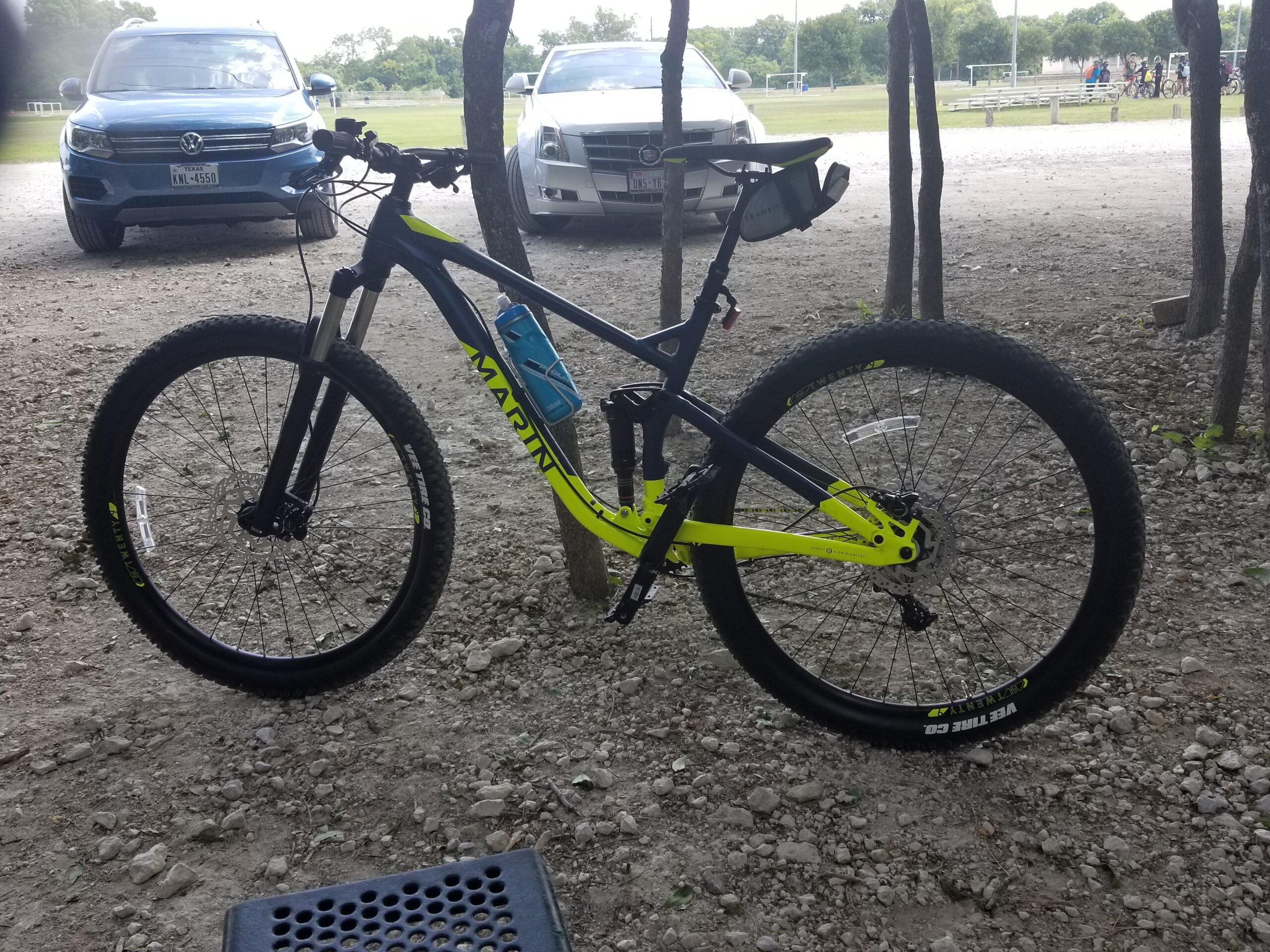 Marin Rift Zone: A black and yellow mountain bike with thick tires is parked on a gravel surface. In the background, two cars are visible, and there is a grassy field with trees and a soccer field beyond. The bike has a water bottle attached to its frame.