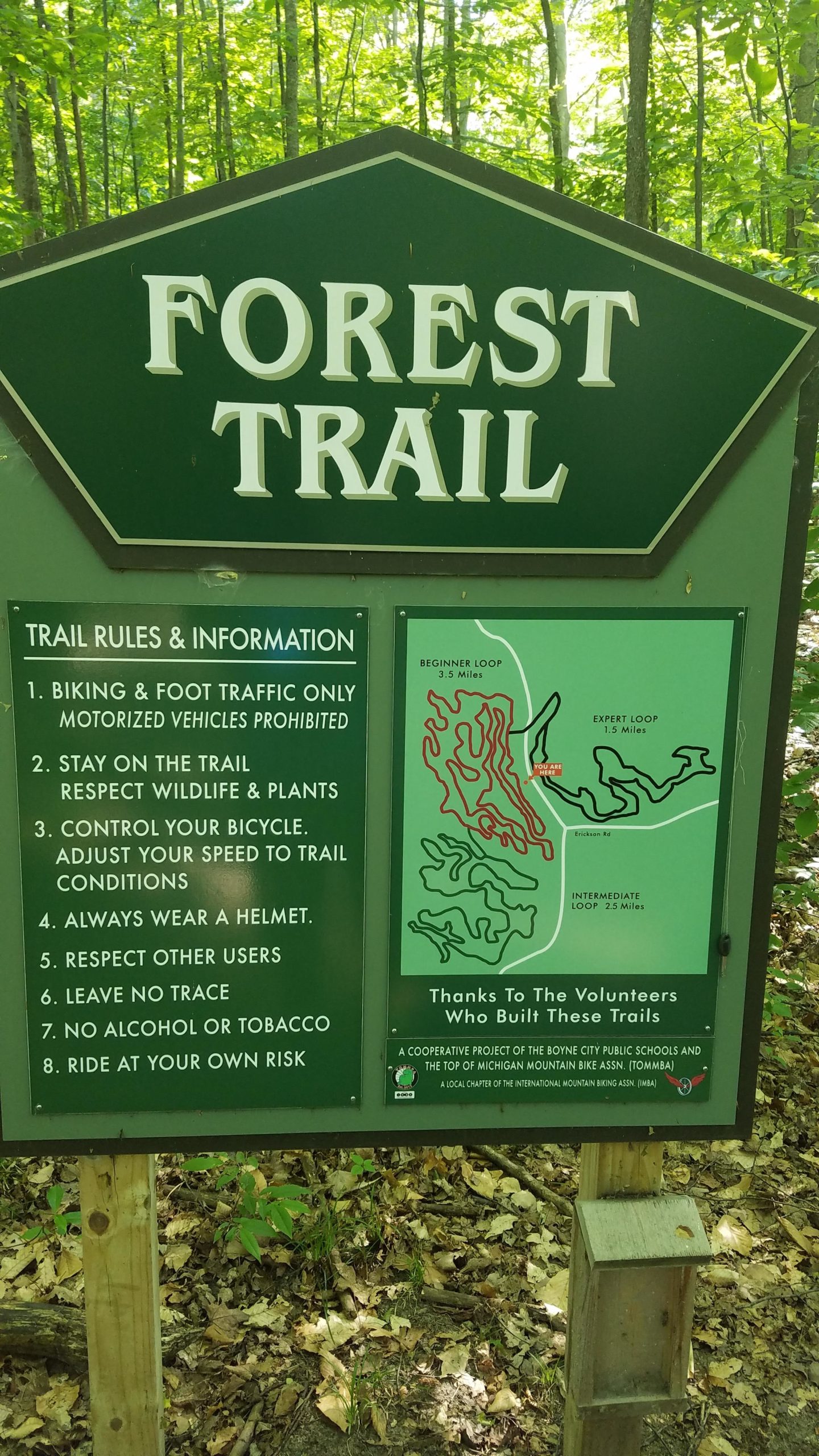 A green sign titled "Forest Trail" displaying trail rules and information, including guidance for biking and foot traffic, safety precautions, and a map outlining beginner, intermediate, and expert loops. The sign is surrounded by a wooded area. Boyne School Forest mountain bike trail.