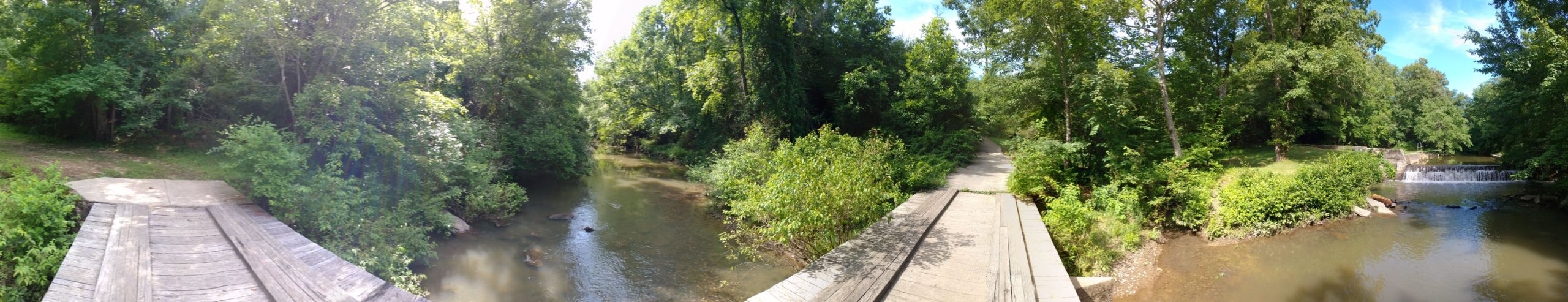 A panoramic view of a serene forested area featuring a wooden bridge leading to a pathway beside a calm creek. Lush green trees and bushes frame the scene, while a small waterfall is visible in the background, enhancing the tranquil ambiance of the natural setting. Piney Creek mountain bike trail.