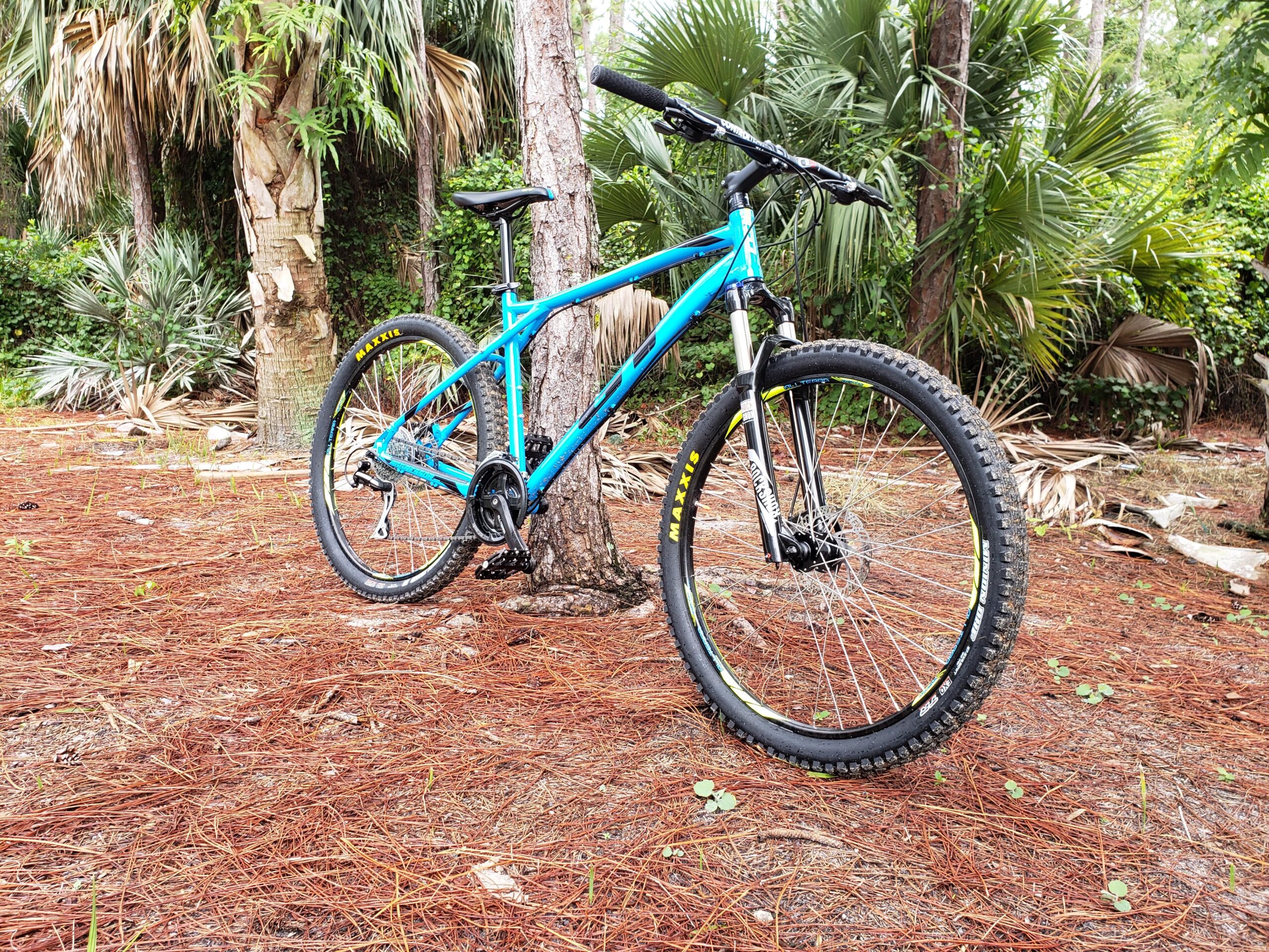 GT Aggressor Pro: A blue mountain bike leaning against a tree in a forested area with palm trees and pine needles on the ground. The bike features thick tires and a sturdy frame, suggesting it's designed for off-road trails.