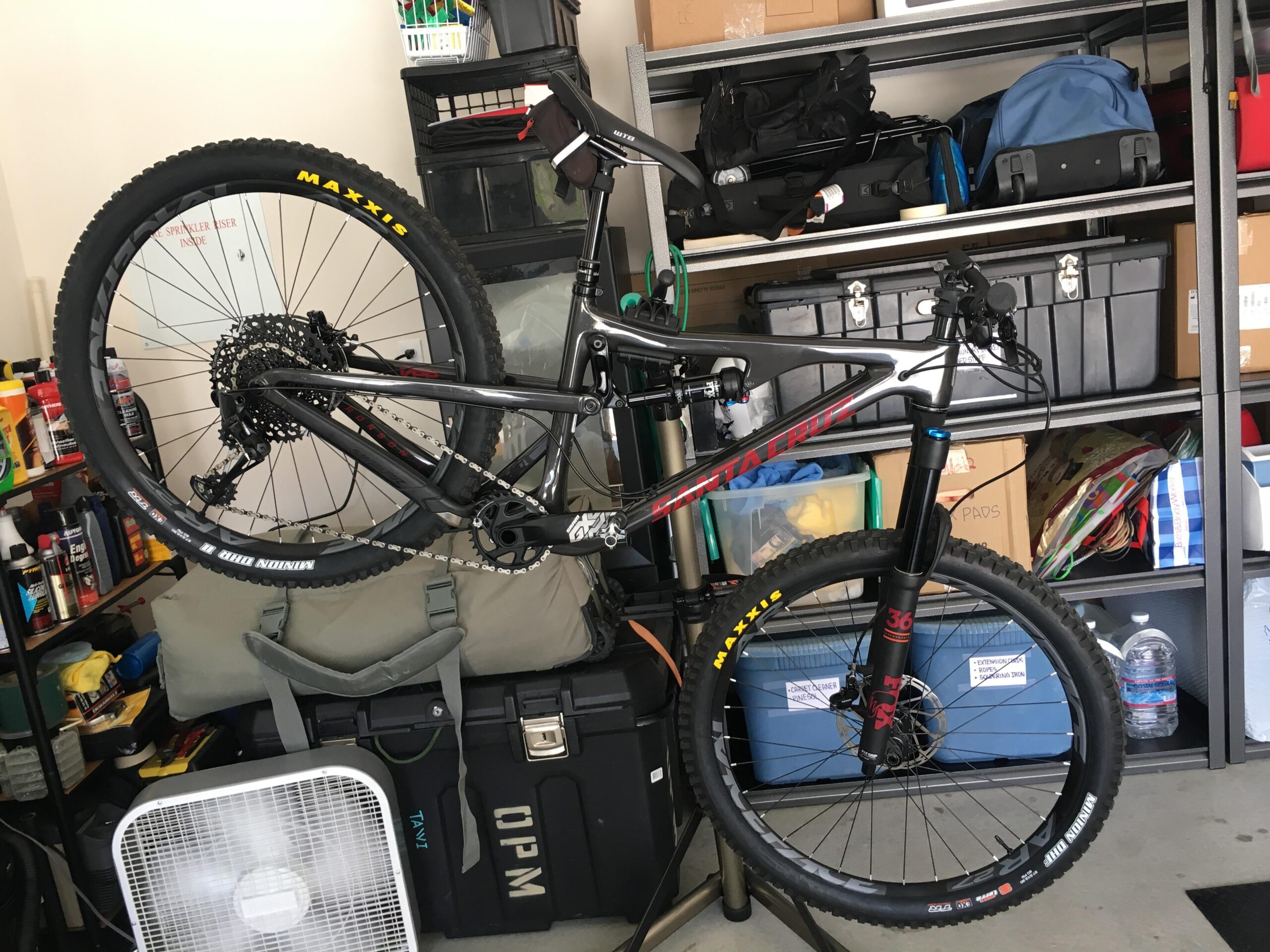 Santa Cruz Bronson: A black mountain bike is displayed upright in a garage, with the front wheel elevated. The bike features Maxxis tires and a prominent Santa Cruz logo. In the background, various storage bins and boxes are visible, along with a fan and several bottles of cleaning products, indicating a cluttered garage environment.