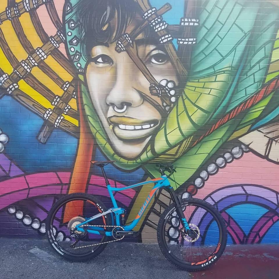Giant Anthem 27.5 2: A colorful urban mural featuring a smiling person's face, adorned with ethnic patterns and accessories, set against a vibrant backdrop. In the foreground, a blue mountain bike is leaning against the wall, complementing the artistic scene.