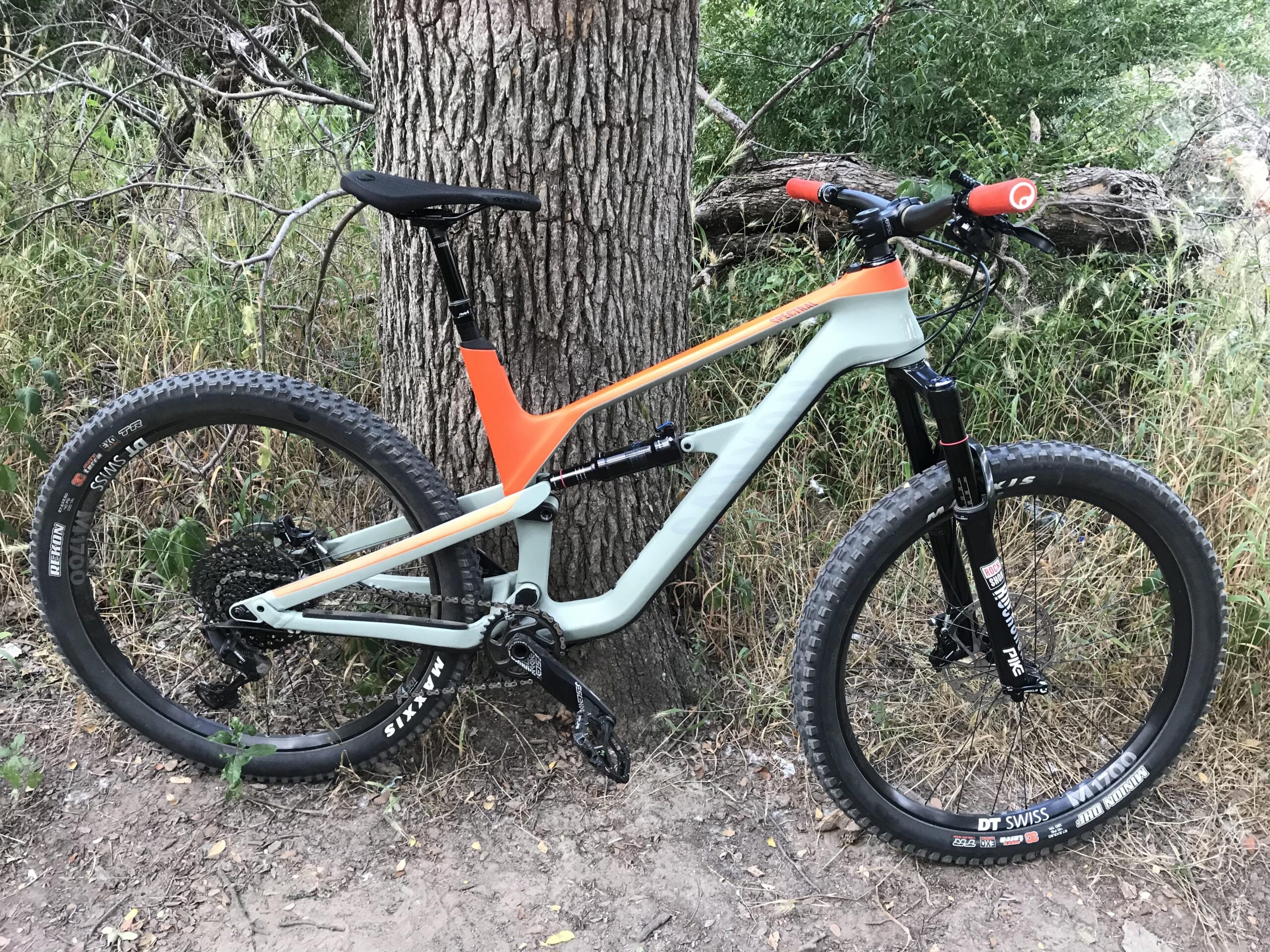 Canyon Spectral CF: A mountain bike with a light green frame and orange accents is leaning against a tree. The bike features thick tires designed for off-road terrain and a visible gear mechanism. Surrounding the bike is a natural setting with tall grass and shrubs.