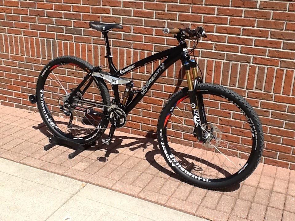 Ellsworth Evolution SST: A black mountain bike with a sleek design and prominent suspension components is leaned against a brick wall. The bike features a silver frame, knobby tires, and is positioned on a paved surface, showcasing its sporty and robust build suitable for off-road riding.