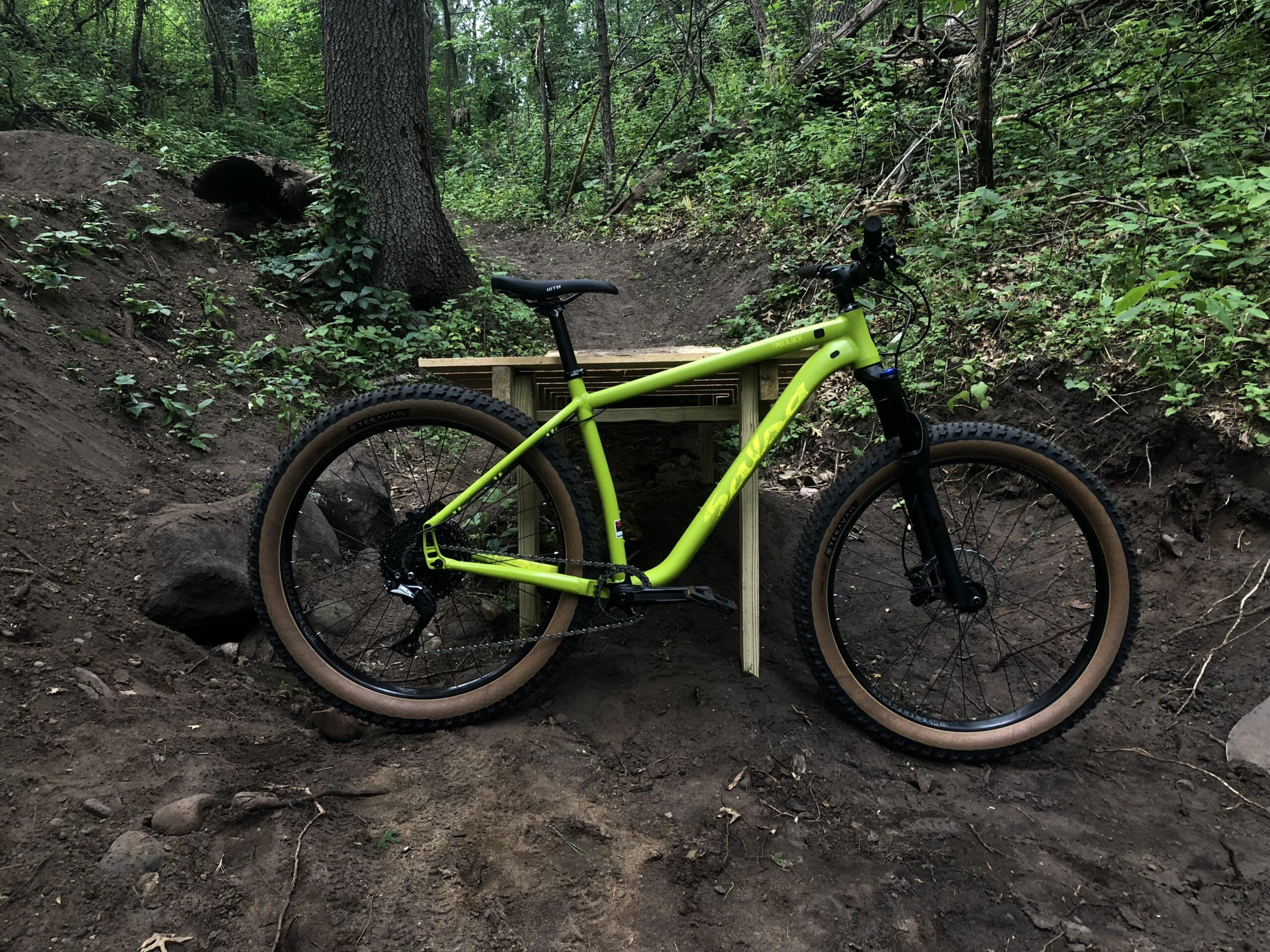 Salsa Timberjack: A bright green mountain bike is positioned next to a wooden jumping ramp in a wooded area. The bike features wide tires with a rugged tread, and the surrounding environment is lush with greenery and dirt, indicating a trail or biking area.