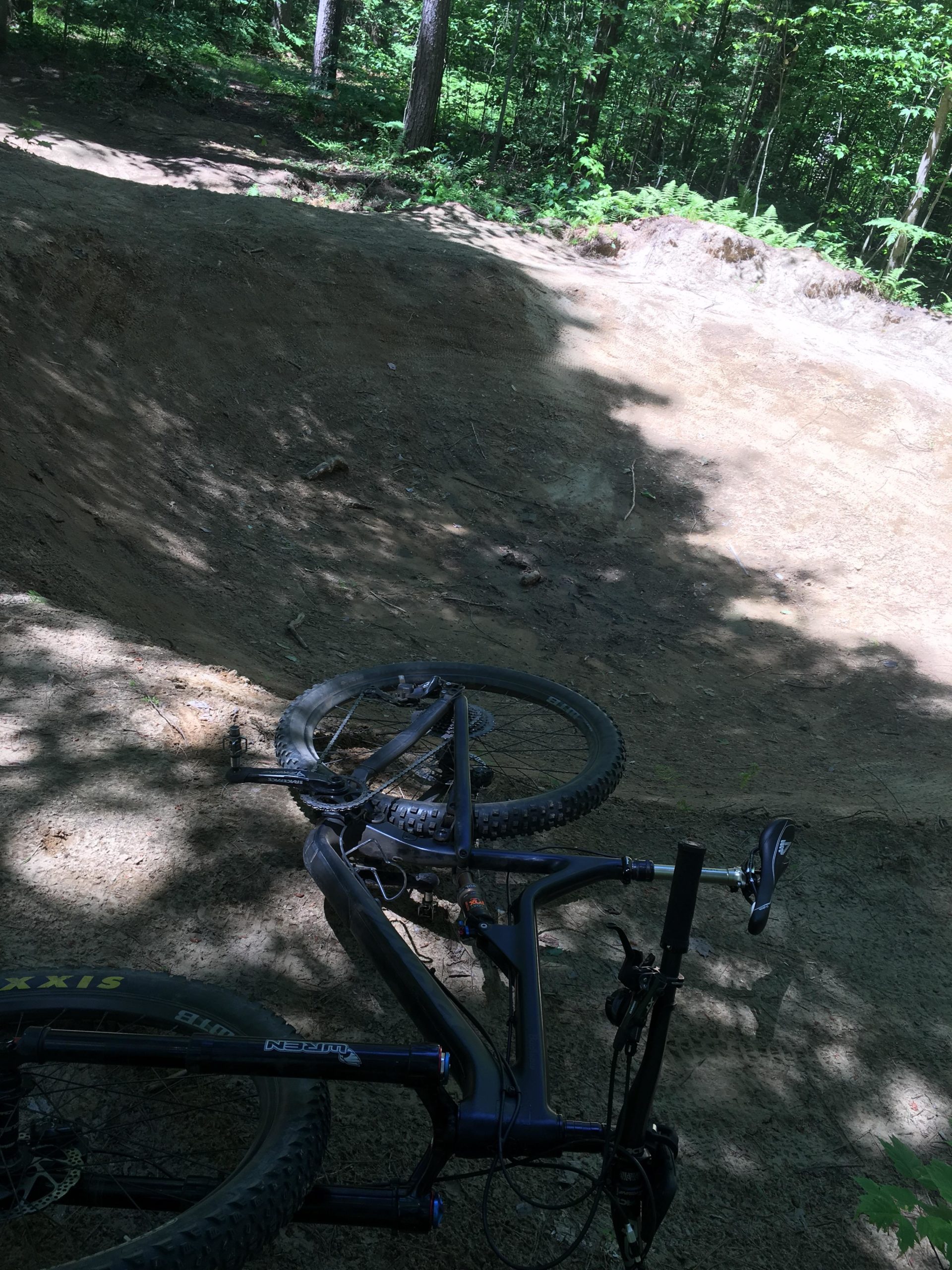 A close-up view of a mountain bike resting on the ground near a dirt jump in a forested area. The bike is positioned on its side, with shafts of sunlight filtering through the trees, highlighting the natural surroundings. The jump is composed of packed dirt and is surrounded by lush greenery. Larose Forest mountain bike trail.