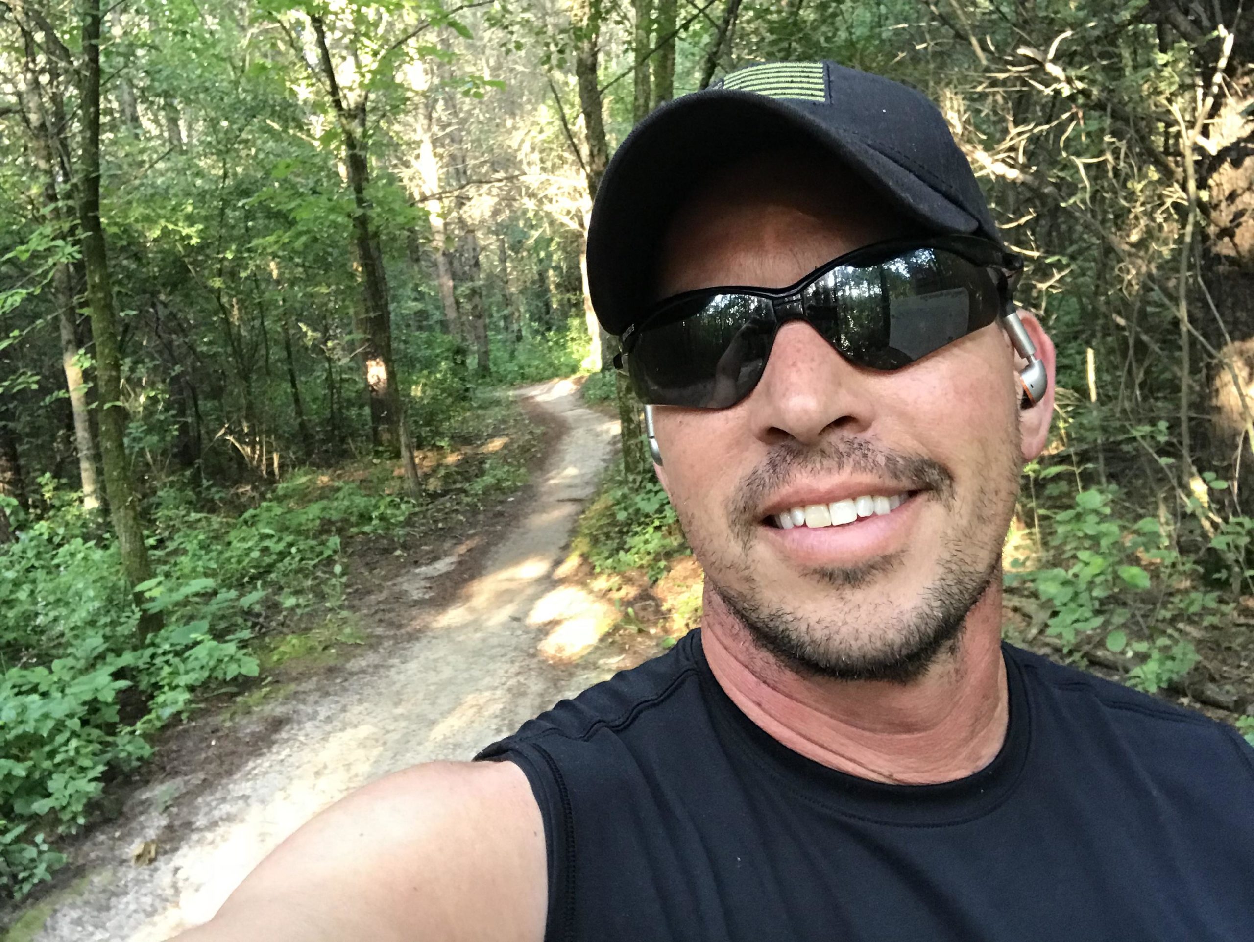 A person wearing sunglasses and a black sleeveless shirt smiles while taking a selfie on a winding trail surrounded by lush green foliage in a wooded area. Lebanon Hills mountain bike trail.