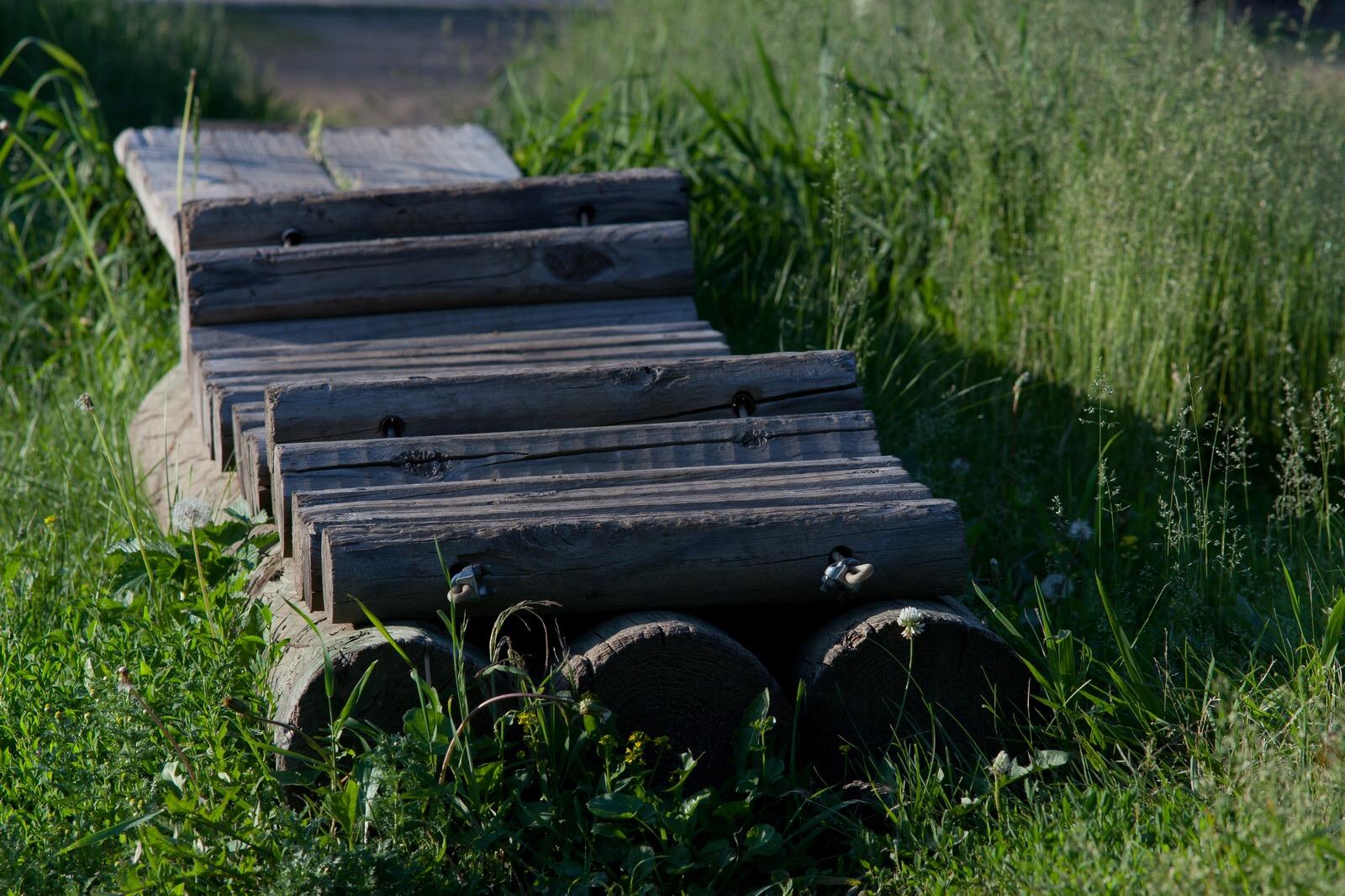 A wooden bridge or path made of logs, partially covered by grass and wildflowers, situated in a natural green setting. Lebanon Hills mountain bike trail.