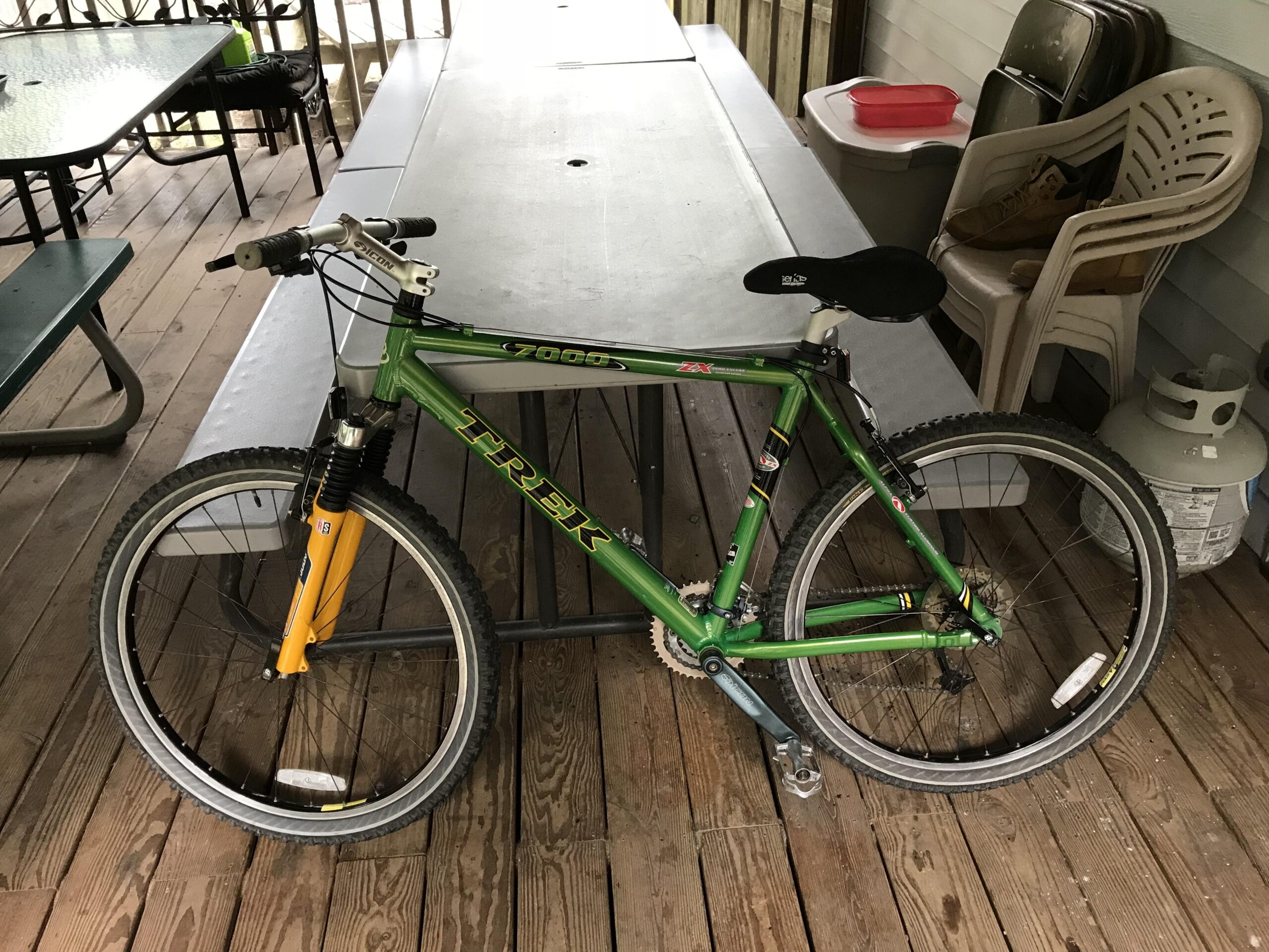 A green Trek mountain bike with yellow front forks is leaning against a picnic table on a wooden deck. Behind the bike, there are plastic chairs stacked, a propane tank, and a small container. The setting appears to be outdoors, possibly at a cabin or a recreational area.
