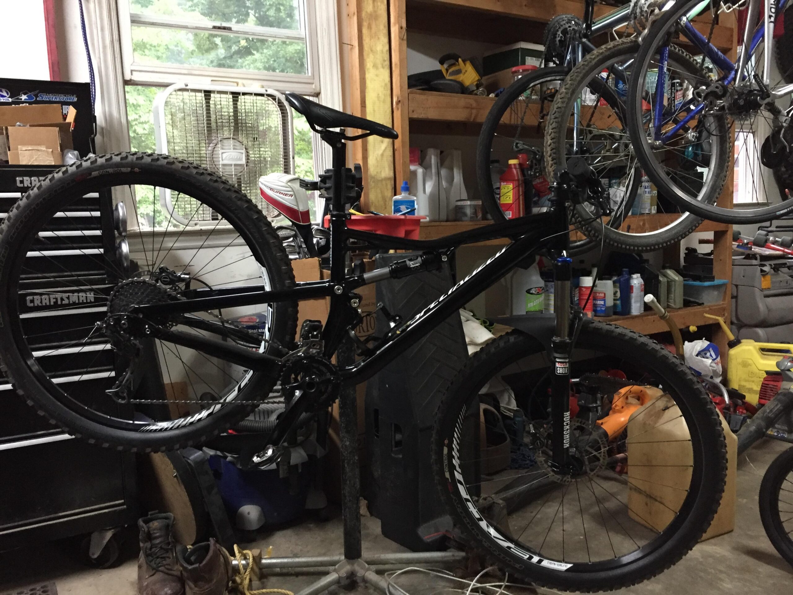 Specialized Camber: A mountain bike is displayed upright in a cluttered garage, with various tools and equipment visible on shelves and the floor. A large window can be seen in the background, along with a fan and multiple bicycles hanging from the ceiling.