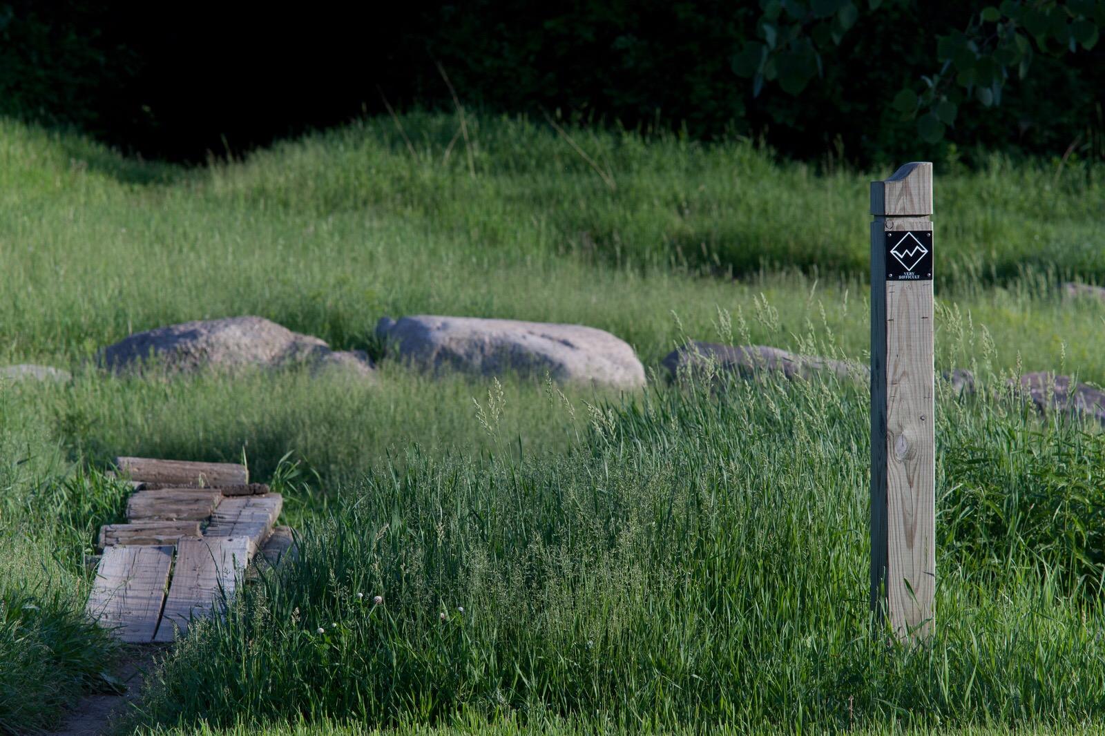 A wooden post with a black and white sign stands in a lush, green area, with tall grass surrounding it. In the foreground, a narrow wooden pathway leads over a small area of grass and rocks, suggesting a natural trail or walking route. Lebanon Hills mountain bike trail.