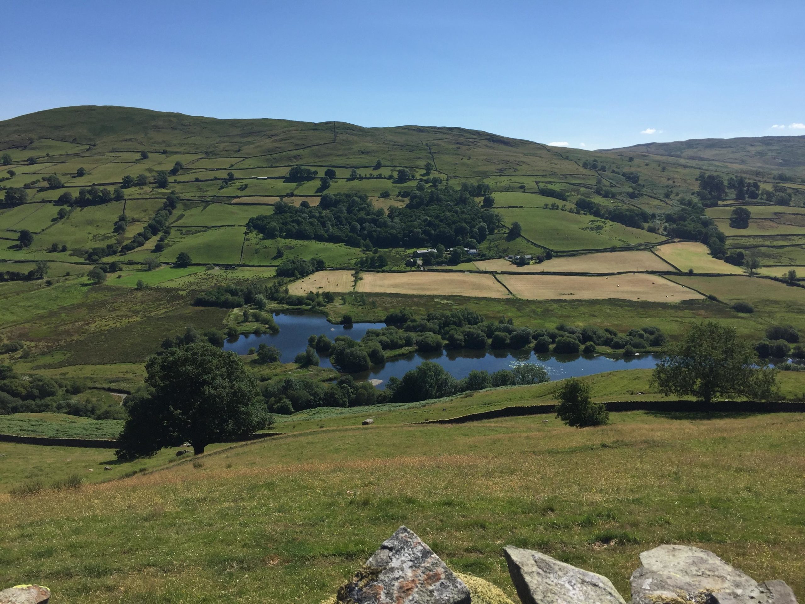 A scenic view of rolling green hills and fields under a clear blue sky, with a tranquil river winding through the landscape. Lush trees dot the hillside, and patches of farmland are visible in the foreground, showcasing a vibrant mix of greenery and earthy tones. Sam Houghton Circuit mountain bike trail.