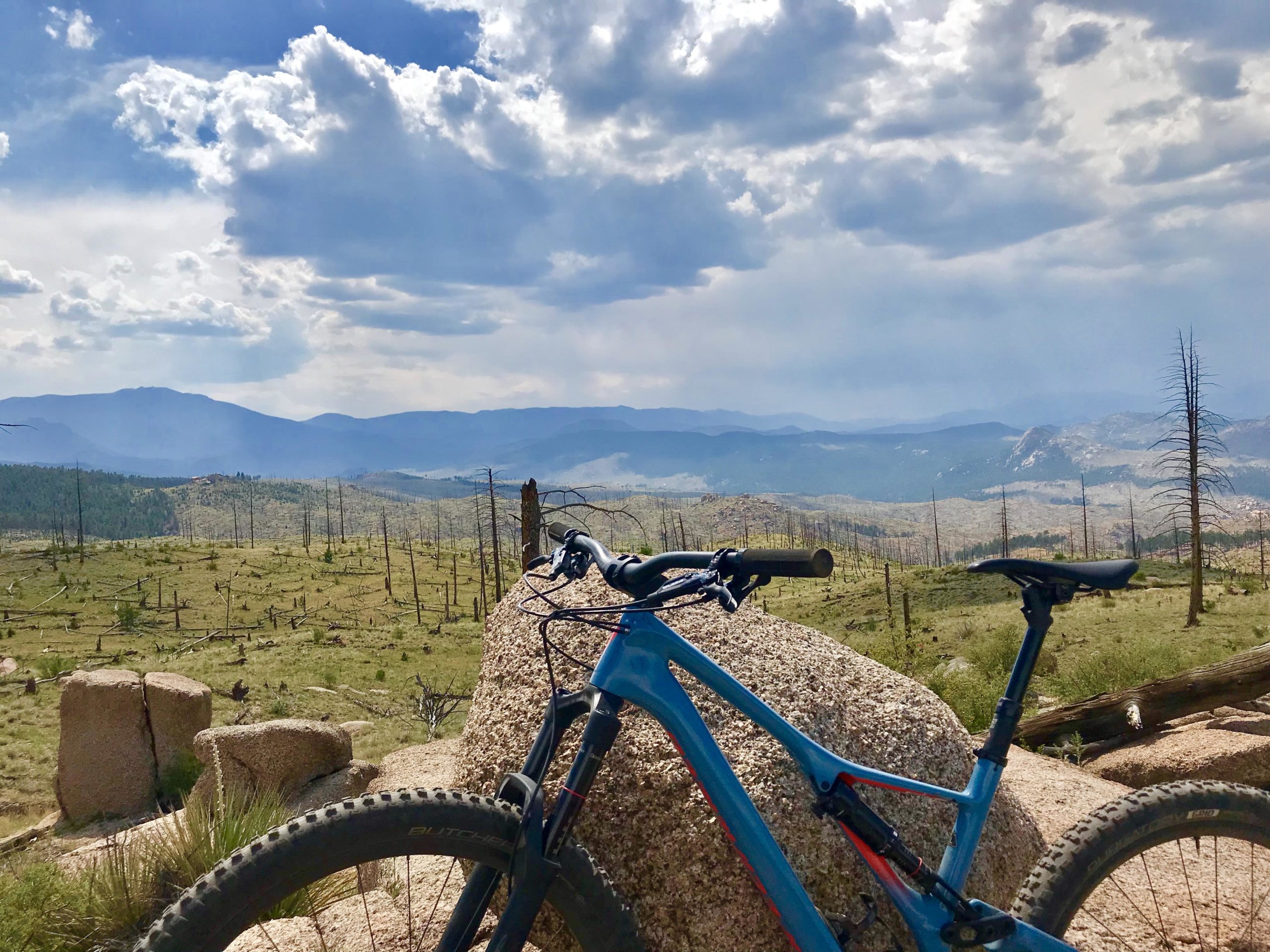 A mountain bike resting on a large rock, overlooking a vast landscape of rolling hills and sparse trees under a partly cloudy sky. The distant mountains create a scenic backdrop, highlighting the natural beauty of the area. Buffalo Creek mountain bike trail.
