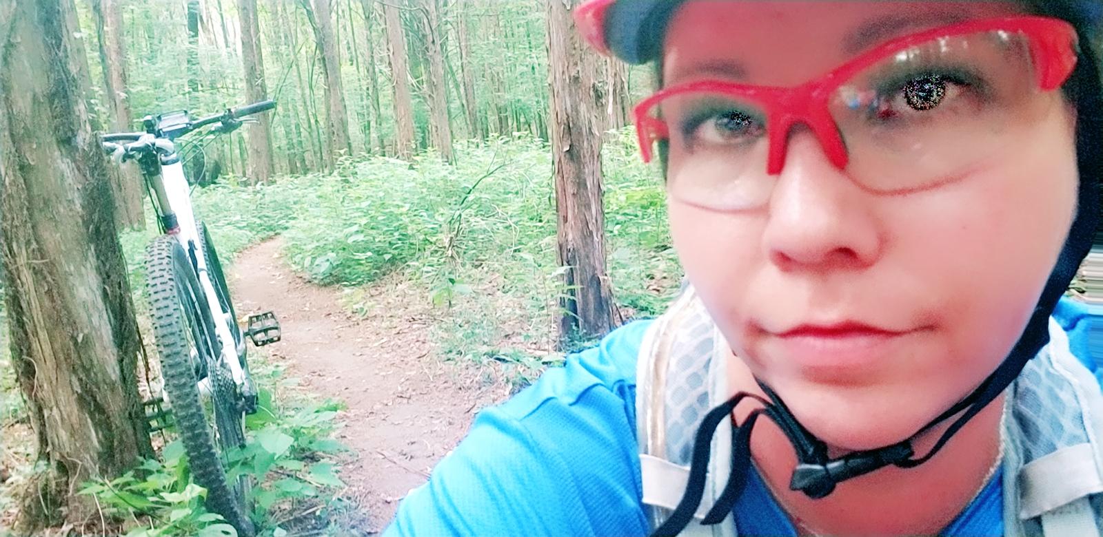 Cannondale Scalpel 29er 3: A person wearing red glasses and a helmet is taking a selfie on a forest trail, with a mountain bike leaning against a tree nearby. Lush greenery surrounds them, and a dirt path can be seen leading deeper into the woods.
