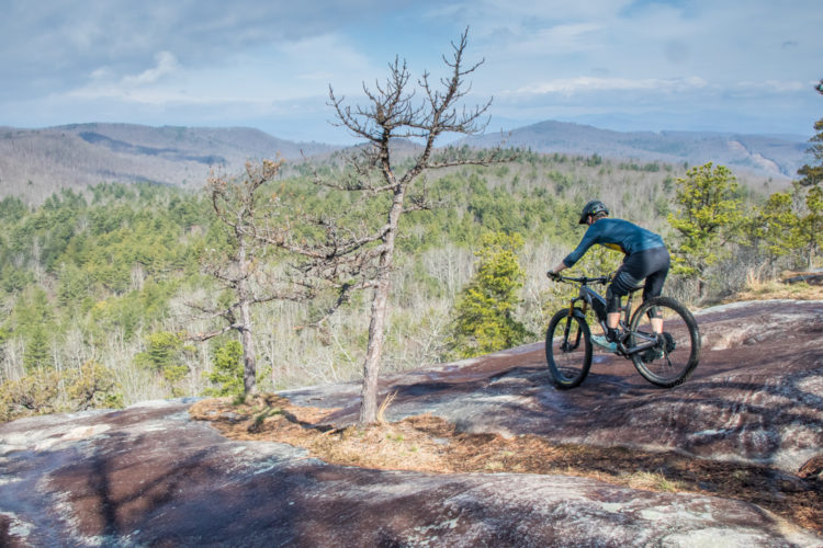 A mountain biker navigating a rocky trail on a hillside overlooking a lush, green valley with distant mountains under a partly cloudy sky.