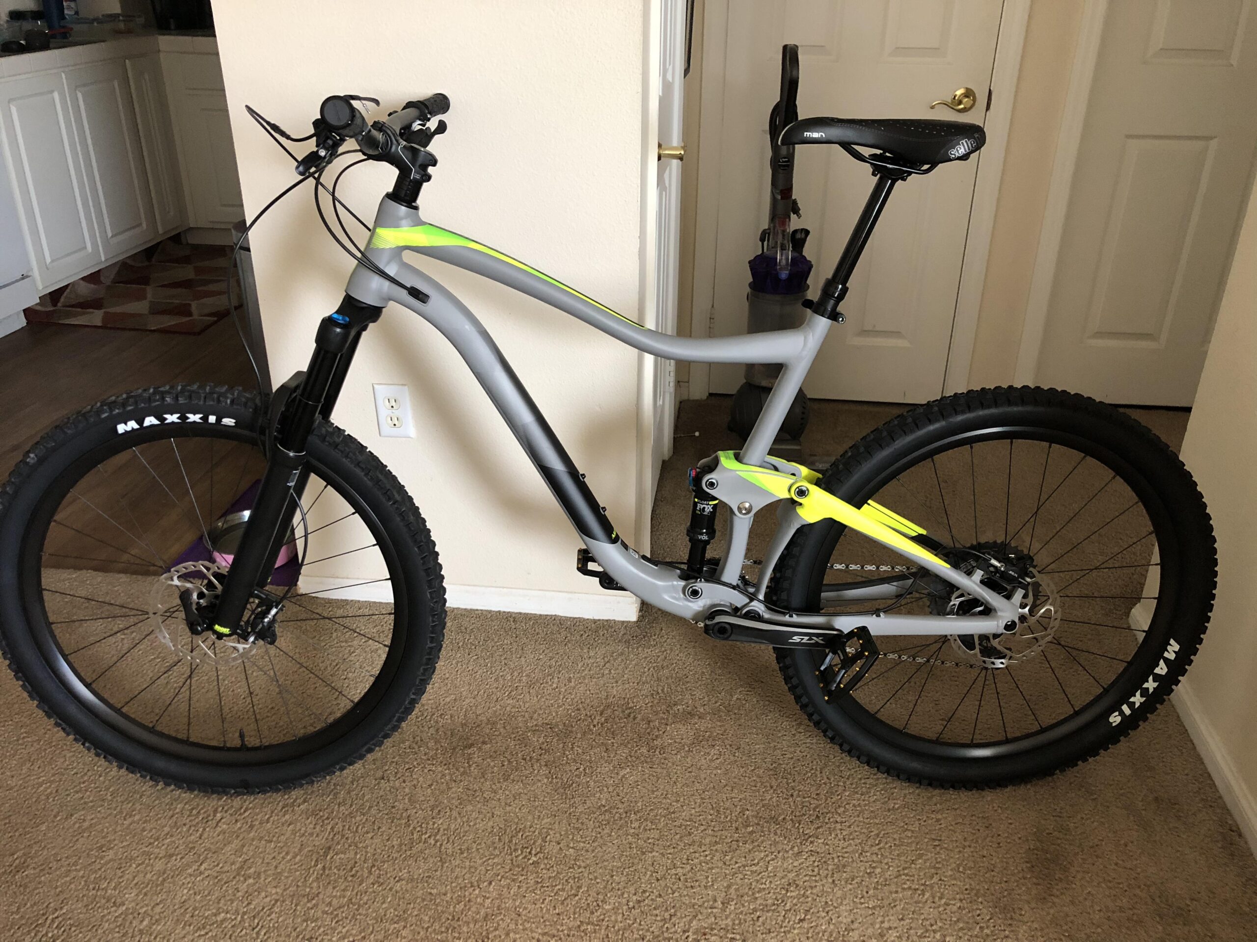 Giant Giant Trance 27.5 2: A modern mountain bike with a gray frame and bright yellow accents, positioned indoors. The bike features wide tires with "Maxxis" branding, a front suspension fork, and disc brakes. In the background, a vacuum cleaner is visible, along with a doorway leading to another room. The floor is carpeted, and there are kitchen cabinets in the distance.