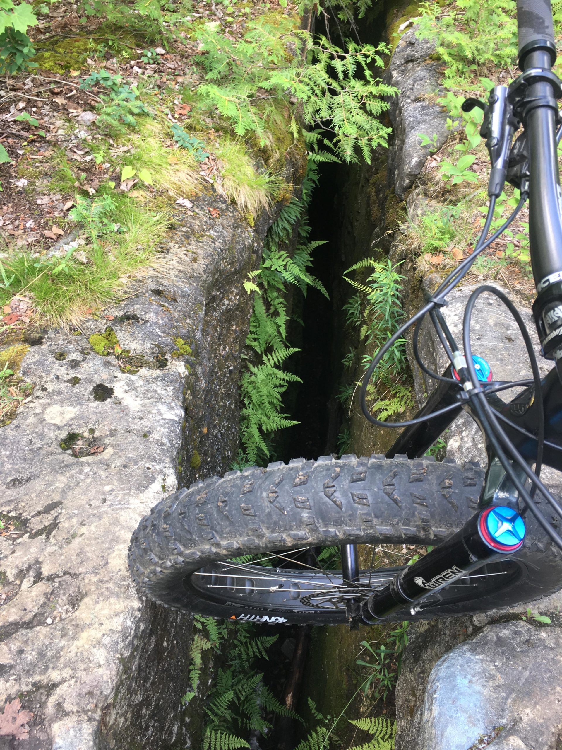 A close-up view of a mountain bike positioned near a narrow crevice in a rocky terrain, surrounded by green ferns and moss. The black tire of the bike hovers above the deep gap, showcasing the steepness of the drop into the shadowy recess below. Inglis falls West rock mountain bike trail.