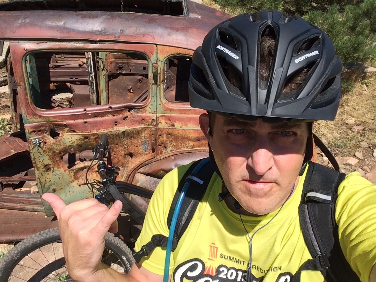 A person wearing a black biking helmet and a bright yellow shirt poses with one hand making a shaka sign. In the background, there is an old, rusted vehicle with a green and red color scheme, partially covered in vegetation. A mountain bike is seen leaning next to the vehicle on a rocky terrain. Wapiti Trail mountain bike trail.
