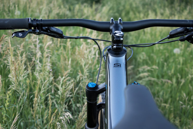 A close-up view of a mountain bike's handlebars and frame, set against a backdrop of tall green grass. The handlebars feature grips and brake levers, while the bike frame showcases a shiny silver finish with a blue knob on the suspension fork.