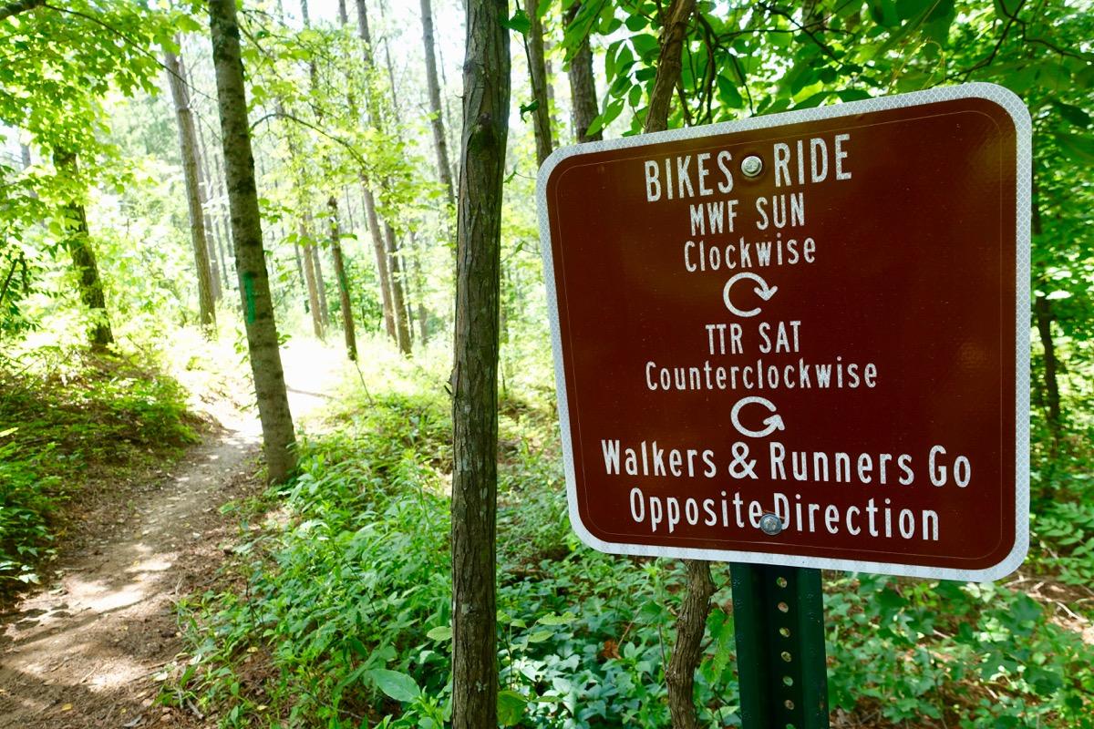 A brown sign in a wooded area providing trail usage directions. The sign states that bicycles should ride clockwise on Mondays, Wednesdays, Fridays, and Sundays, while they should ride counterclockwise on Tuesdays, Thursdays, and Saturdays. It also informs walkers and runners to go in the opposite direction. Sunlight filters through the trees, illuminating the path ahead. Talking Rock Nature Preserve mountain bike trail.