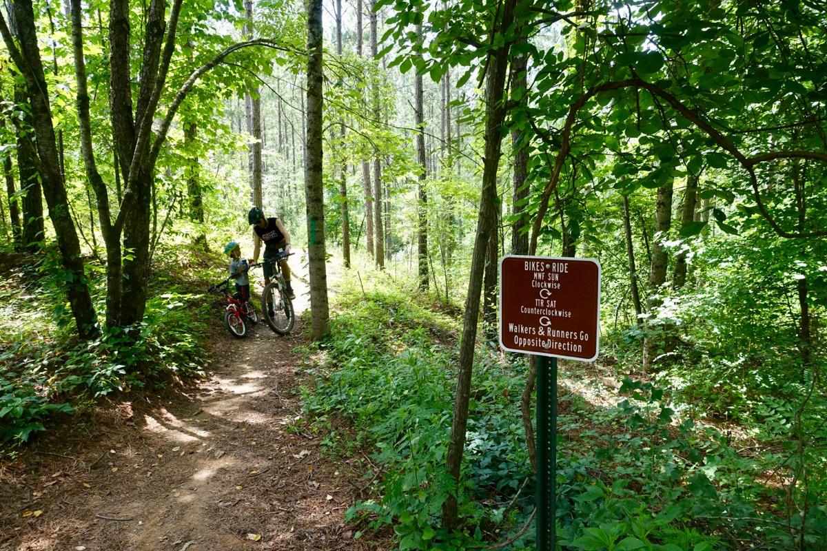 A child and an adult wearing helmets stand beside a bicycle on a narrow dirt trail in a wooded area, surrounded by lush green foliage. A sign indicates trail usage rules for bikes and walkers. Sunlight filters through the trees, creating a bright and inviting atmosphere. Talking Rock Nature Preserve mountain bike trail.