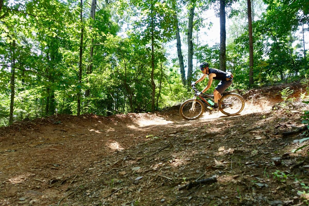 A mountain biker navigating a dirt trail in a forested area, surrounded by lush green trees and sunlight filtering through the leaves. The biker is leaning into a curve, showcasing an active outdoor lifestyle. Talking Rock Nature Preserve mountain bike trail.