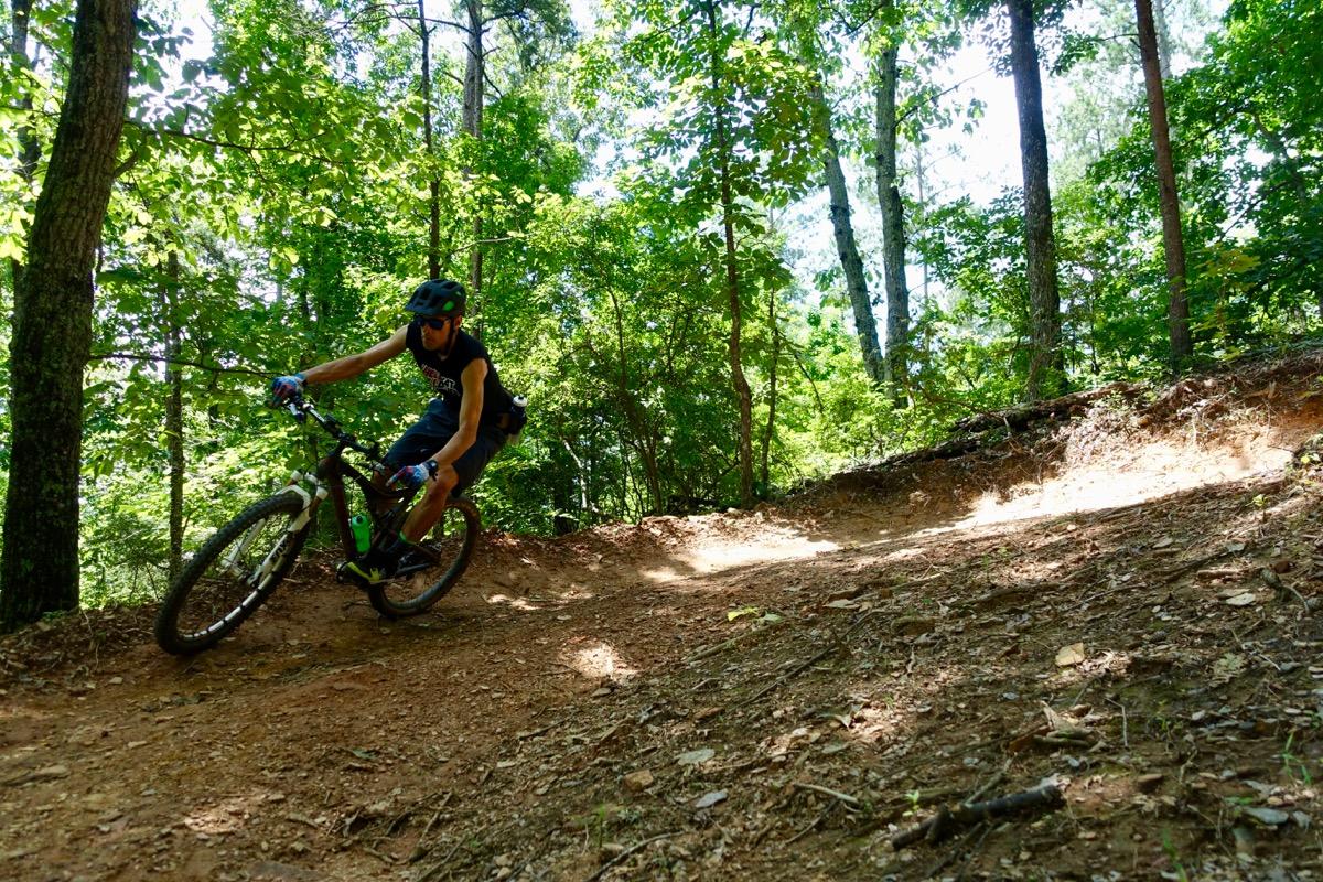 A person riding a mountain bike on a dirt trail in a dense forest, leaning into a turn with trees and greenery surrounding the path. The sun filters through the leaves, creating a vibrant outdoor scene. Talking Rock Nature Preserve mountain bike trail.