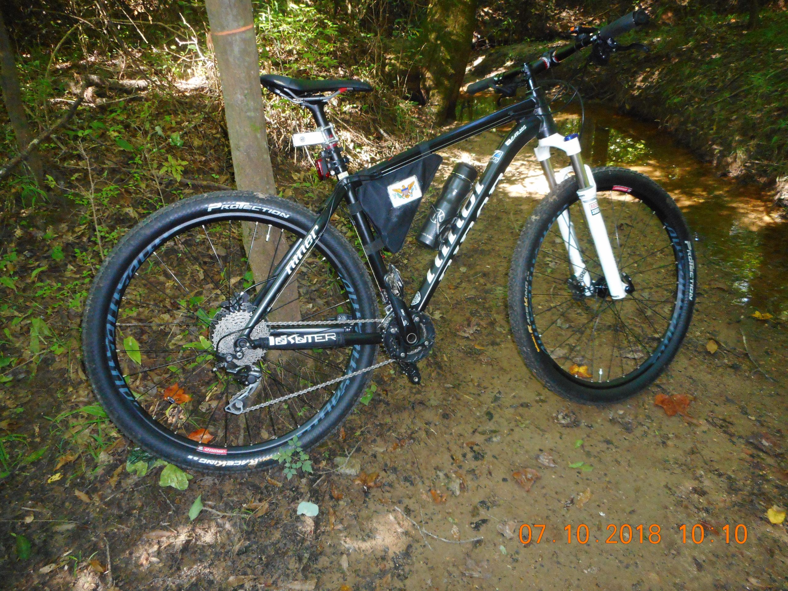 A black mountain bike positioned beside a small stream in a wooded area, with lush green foliage and earthy ground. The bike features a sleek frame, wide tires, and a water bottle attached to the frame. Yellow River mountain bike trail.