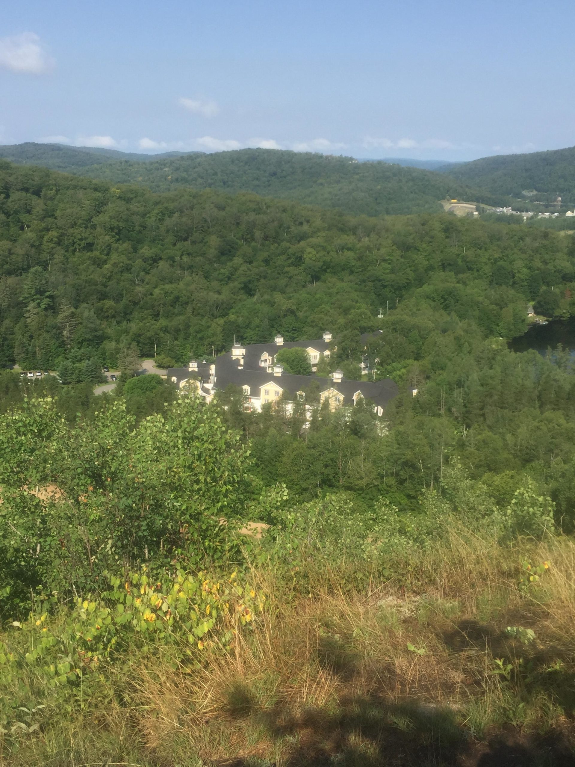 A scenic view of a mountainous landscape featuring lush green trees and a distant building nestled in the valley, under a clear blue sky with a few clouds. Mont-tremblant, Coupe Quebec Dh&xc mountain bike trail.